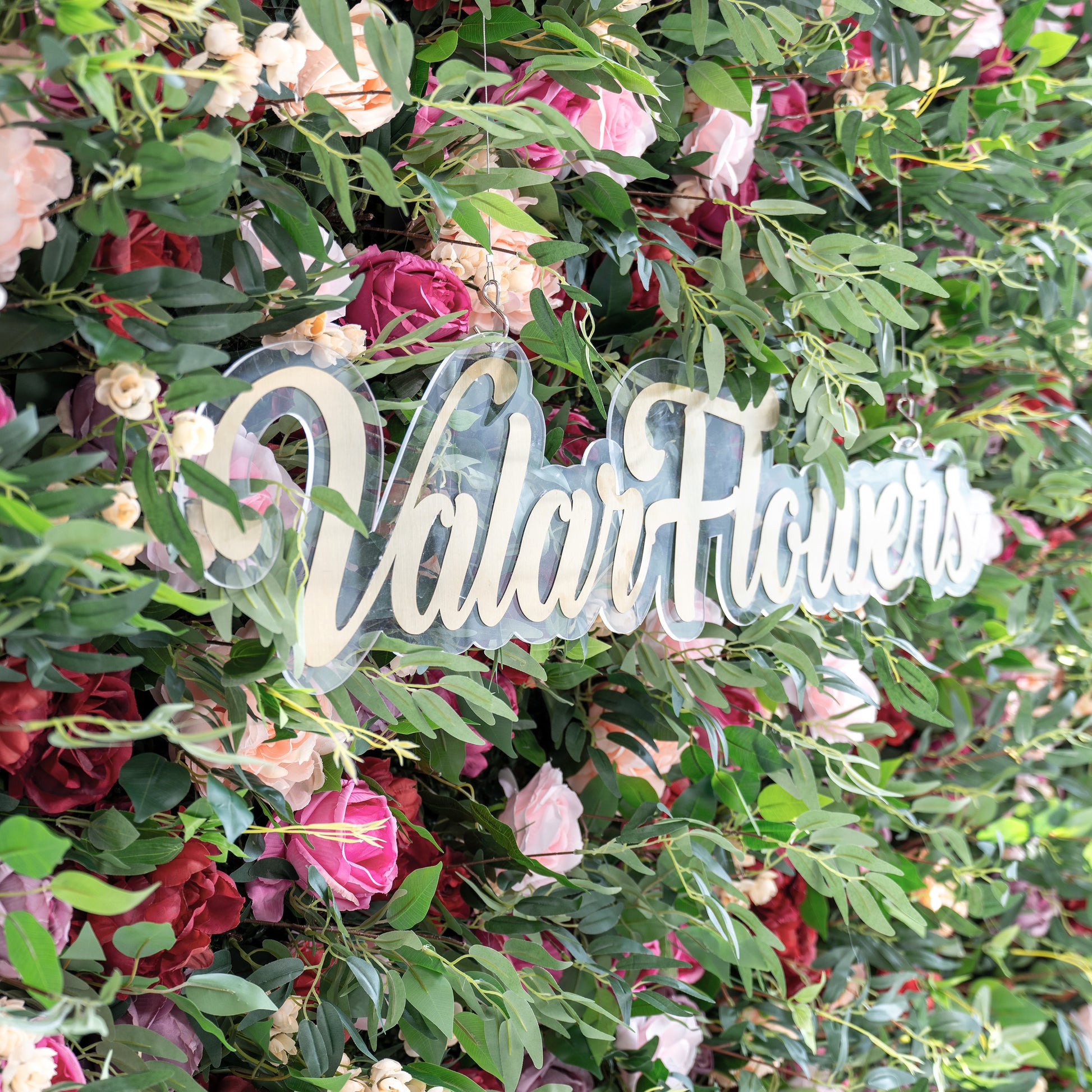 A close - up of a clear acrylic sign with gold “ValarFlowers” script, nestled in a lush artificial flower wall. The wall features pink, red, and peach roses, plus green foliage, creating a vibrant, decorative backdrop that highlights the branding element. 
