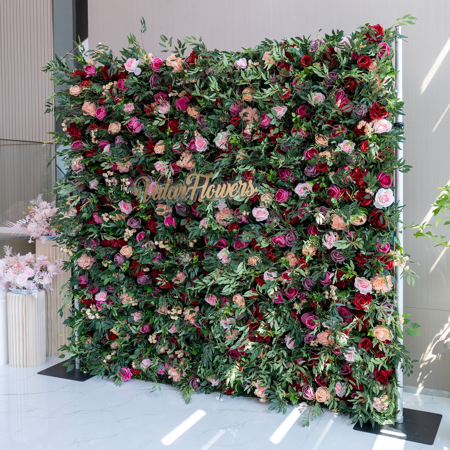A large, lush artificial flower wall features a vibrant mix of roses in pink, red, purple, and peach, plus green foliage and small white flowers. A gold “ValarFlowers” sign stands out in the center. It’s an eye - catching, elegant backdrop, perfect for events or decorative displays. 