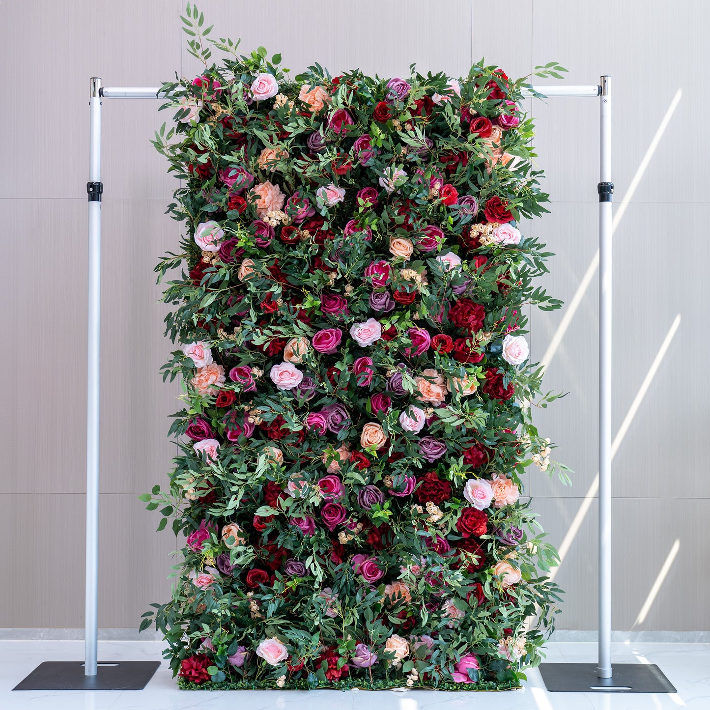A tall, rectangular artificial flower wall features a lush mix of roses in pink, red, purple, and peach, along with green foliage. Mounted on a metal frame, it creates an elegant, vibrant backdrop, perfect for events or decorative displays. 