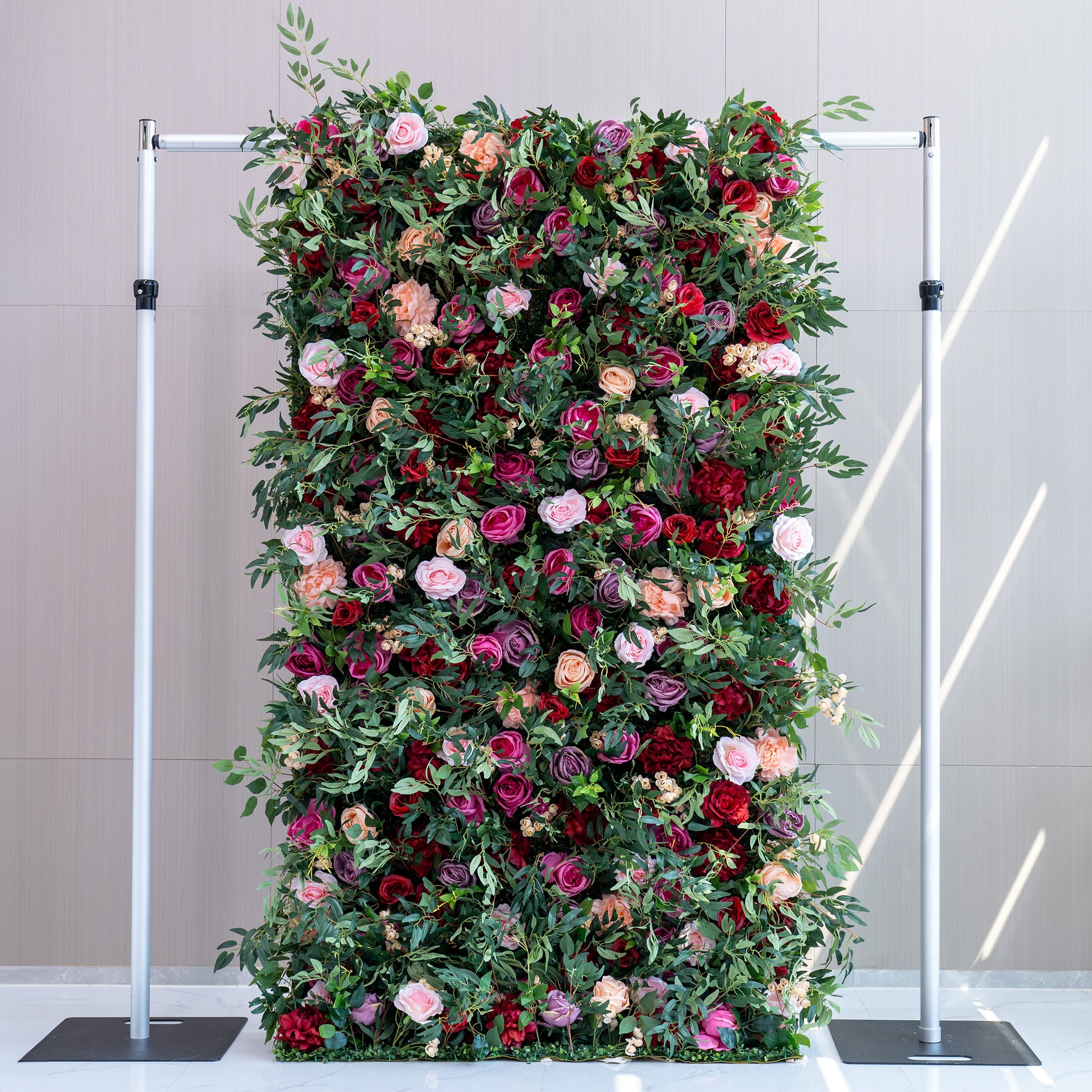 A tall, rectangular artificial flower wall features a lush mix of roses in pink, red, purple, and peach, along with green foliage. Mounted on a metal frame, it creates an elegant, vibrant backdrop, perfect for events or decorative displays. 