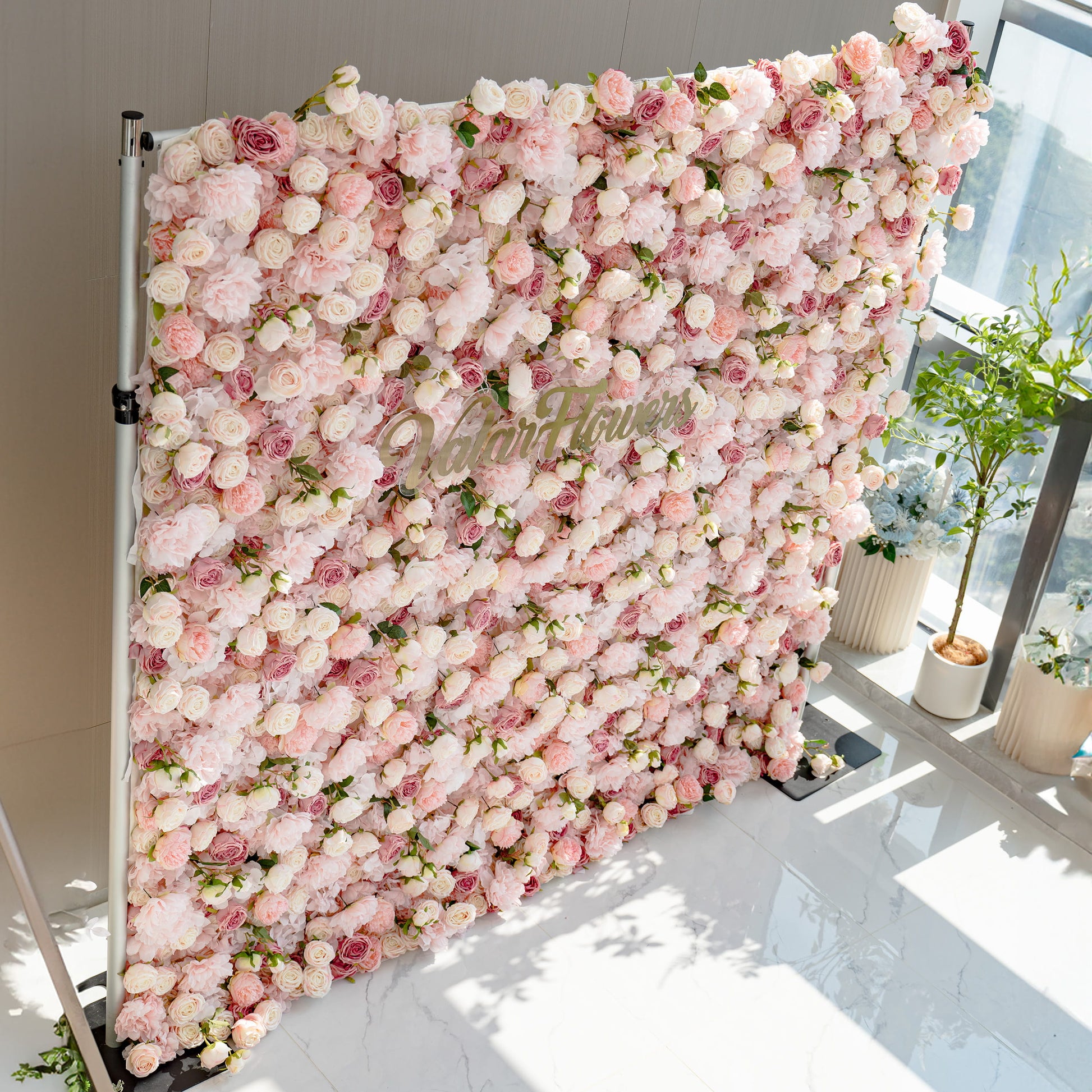 A stunning flower wall filled with a variety of pink and white roses. The wall is labeled "ValarFlowers" and is set against a modern interior with potted plants nearby.