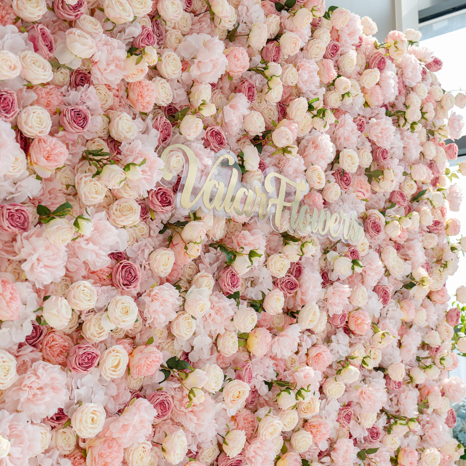A beautiful flower wall composed of various pink and white roses and peonies. The words "Valar Flowers" are prominently displayed in the center, creating a romantic and elegant atmosphere.
