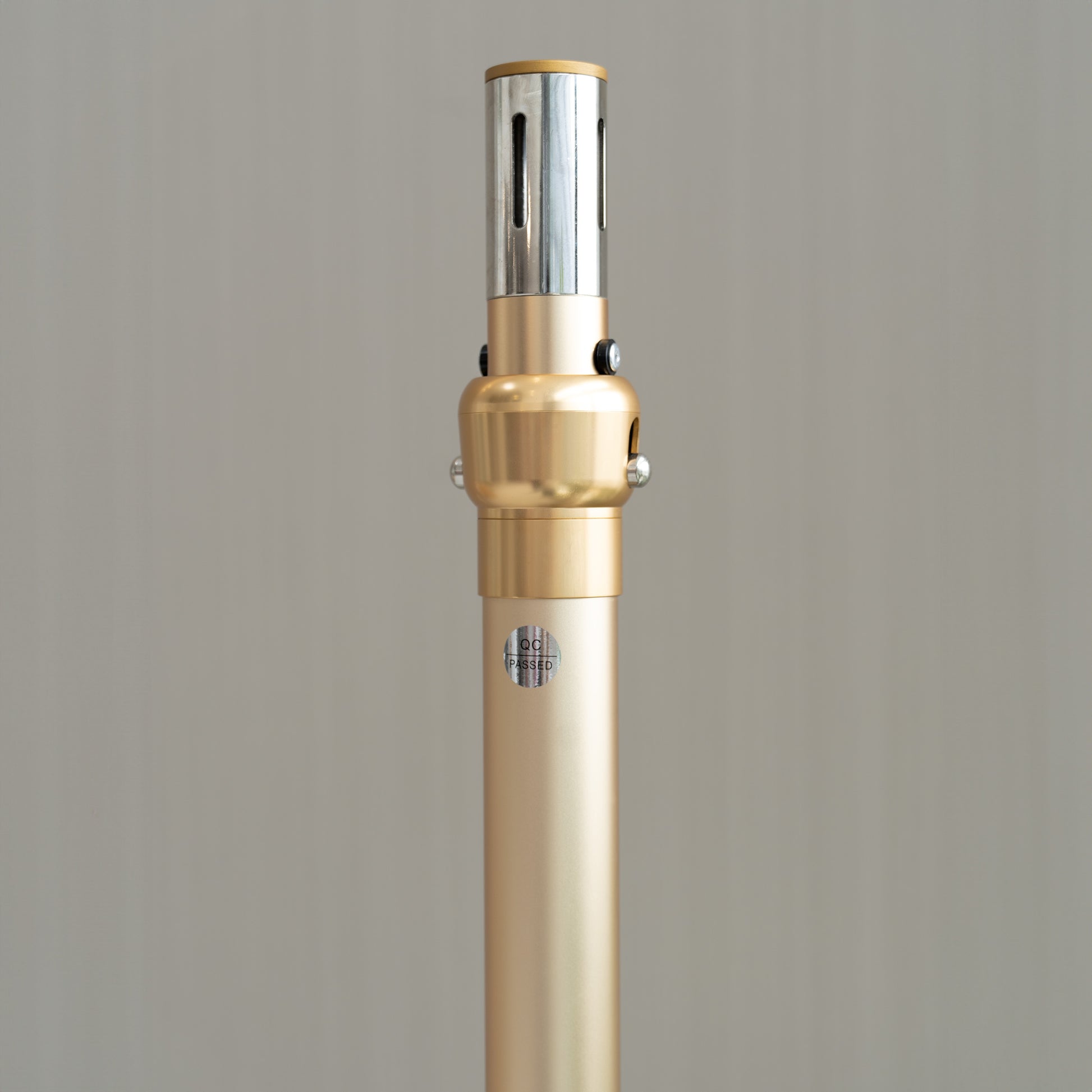 This image showcases a metallic pole with a sophisticated design. The pole is predominantly golden with a sleek, polished finish. At the top, there is a silver - colored section with vertical slits. The middle part features a golden, rounded joint with two metallic knobs, likely for adjustment or connection purposes. A small "QC PASSED" label is affixed to the pole, indicating quality control approval. 