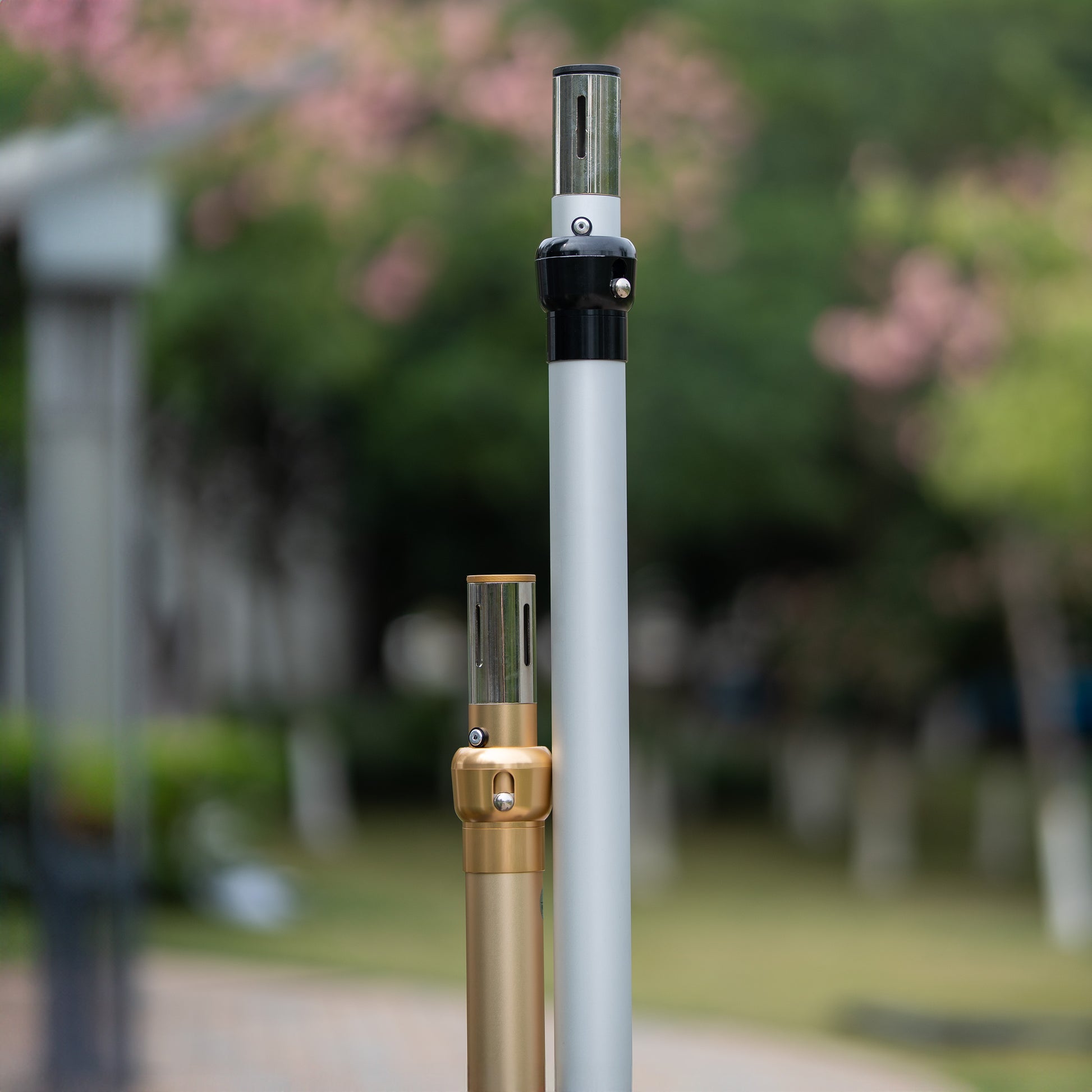In the image, two vertical poles stand against a softly blurred outdoor background with greenery and hints of pink flowers. One pole is golden with a gold - toned connector, while the other is silver - gray with a black connector. Both poles have metallic tops with slots, likely for assembling structures, such as backdrop stands for events or photography. 