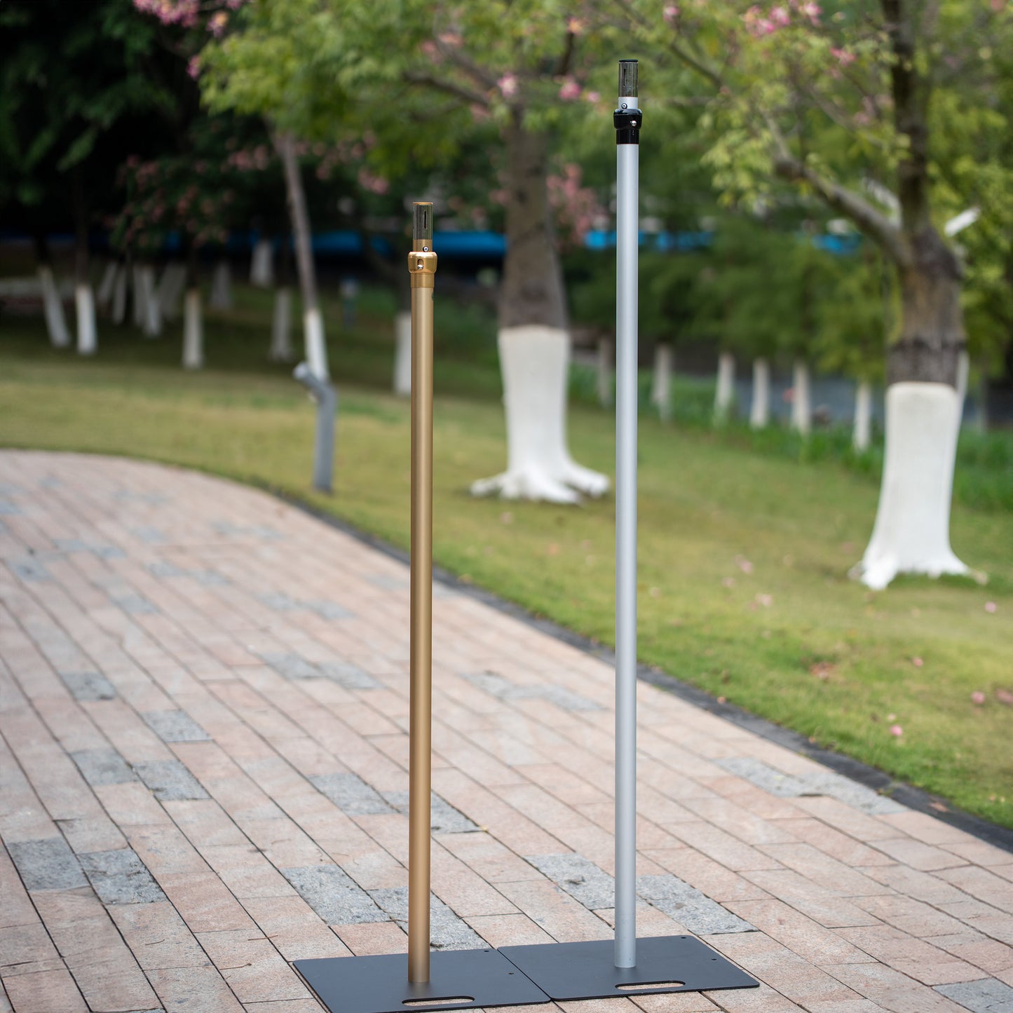 In the image, two vertical poles stand on flat black bases placed on a paved pathway. One pole is gold - colored, and the other is silver - gray. The background features a park - like setting with green grass, trees with white - painted trunks, and some pink flowers. The poles appear to be parts of a structure, possibly for supporting backdrops or decorations, and are designed to be sturdy and stable. 