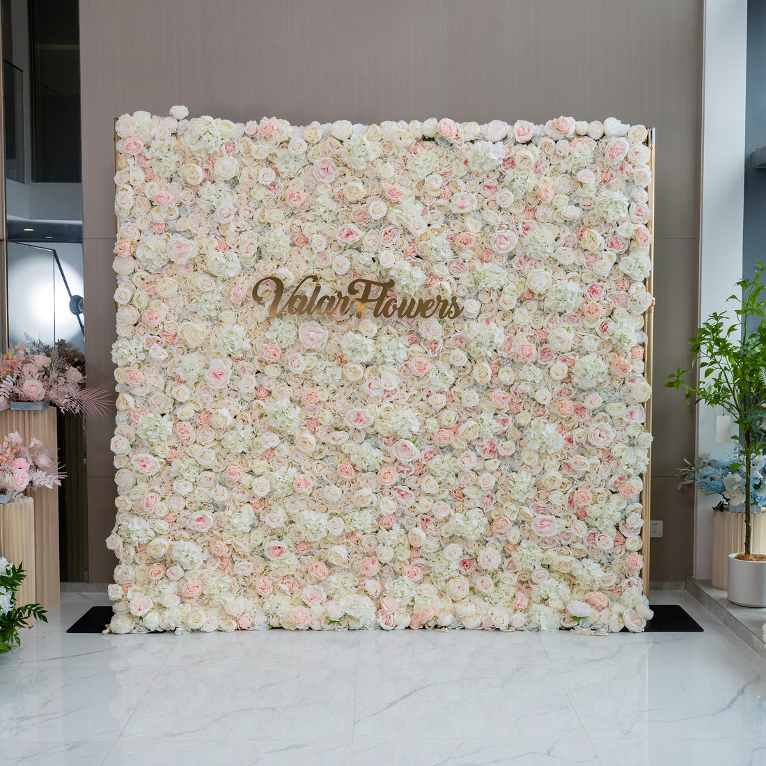 This image showcases a floral wall adorned with a dense arrangement of light pink and white flowers. The wall features the brand name "ValarFlowers" prominently displayed in the center. The overall ambiance is soft, romantic, and inviting, making it ideal for events such as weddings, bridal showers, or romantic celebrations.
