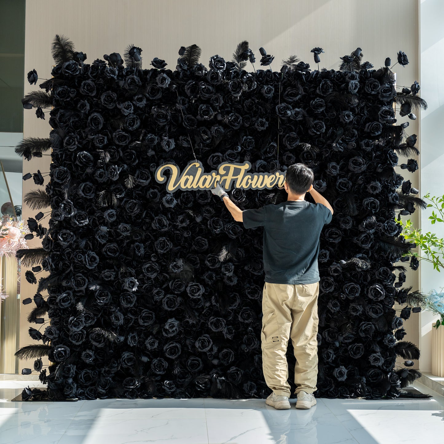 A person is setting up a sign for "ValarFlowers" in front of a stunning black rose wall. The wall, adorned with feathers, creates a dramatic and elegant display.