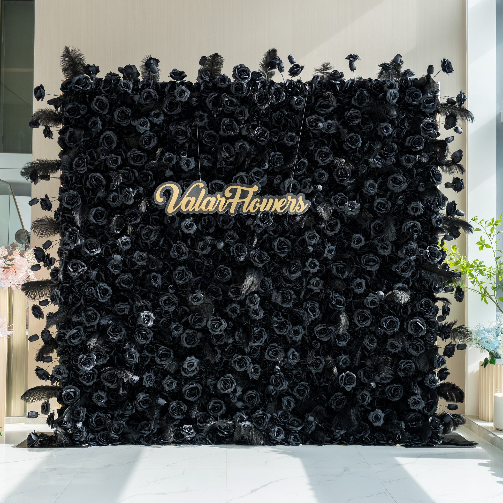 The image features a striking black floral wall with the name "ValarFlowers" prominently displayed in the center. The wall is adorned with black roses and feathers, creating a dramatic and elegant look.
