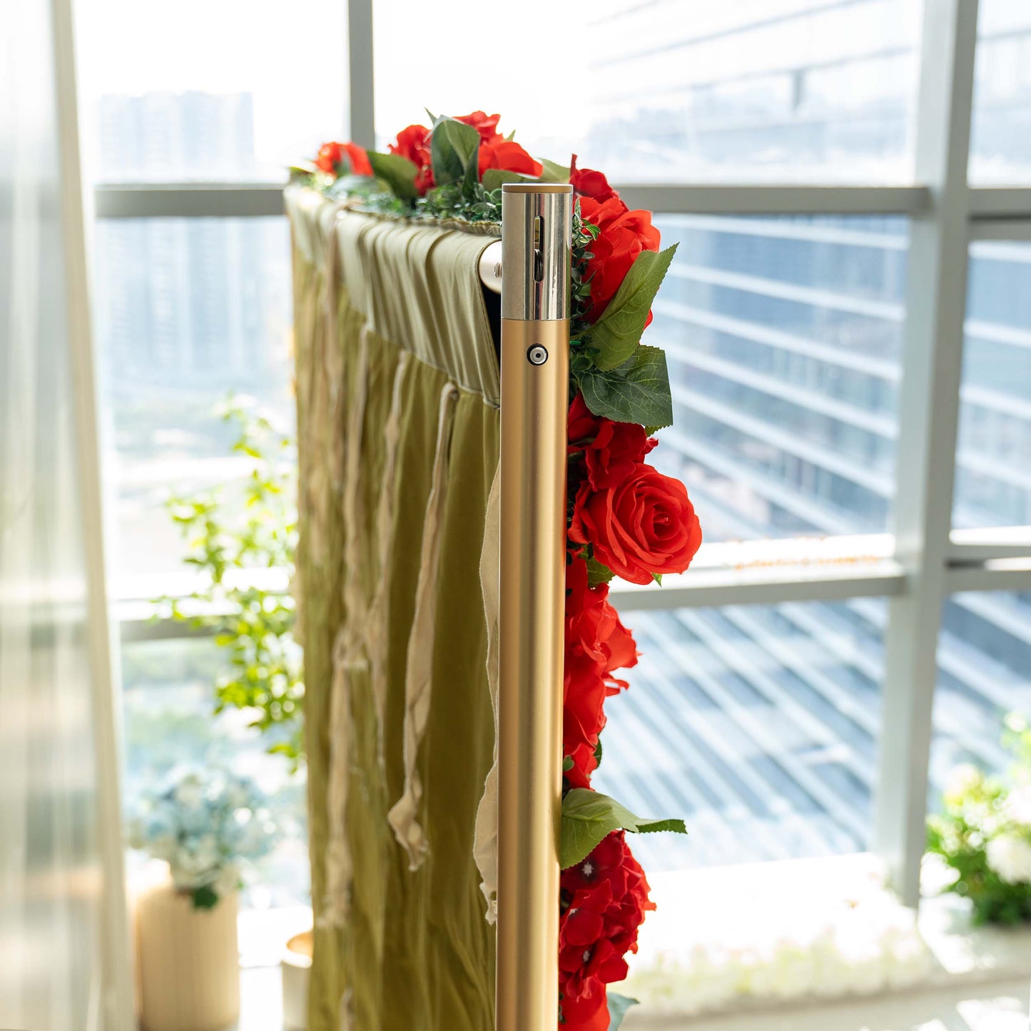 A close - up shows a metallic stand adorned with vibrant red artificial roses and green leaves, with a fabric panel attached, set against a window with a city view. 