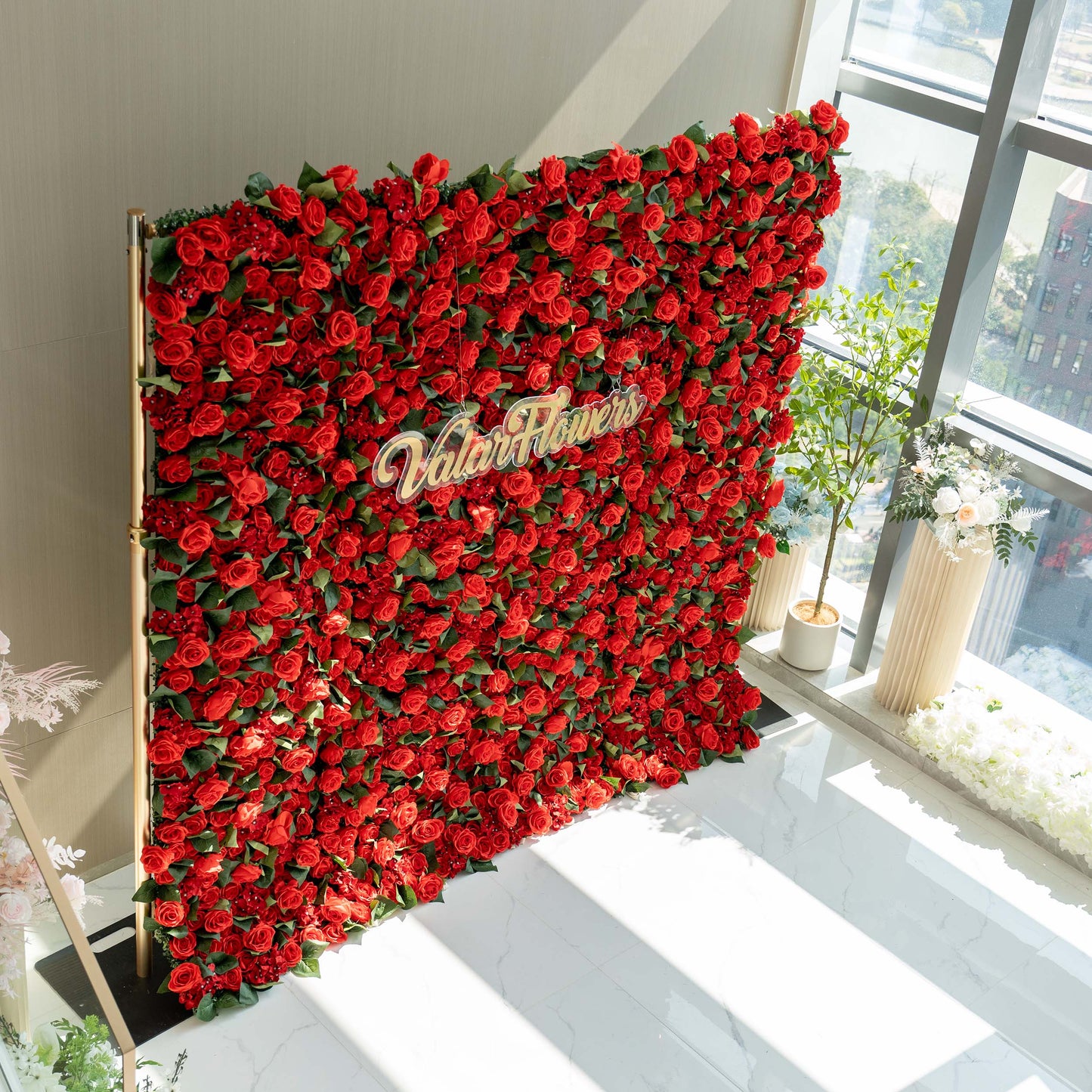 A striking red floral wall by ValarFlowers, densely packed with artificial roses and green leaves. Set near a window, it makes an elegant backdrop with the brand name ValarFlowers aprominently displayed. 