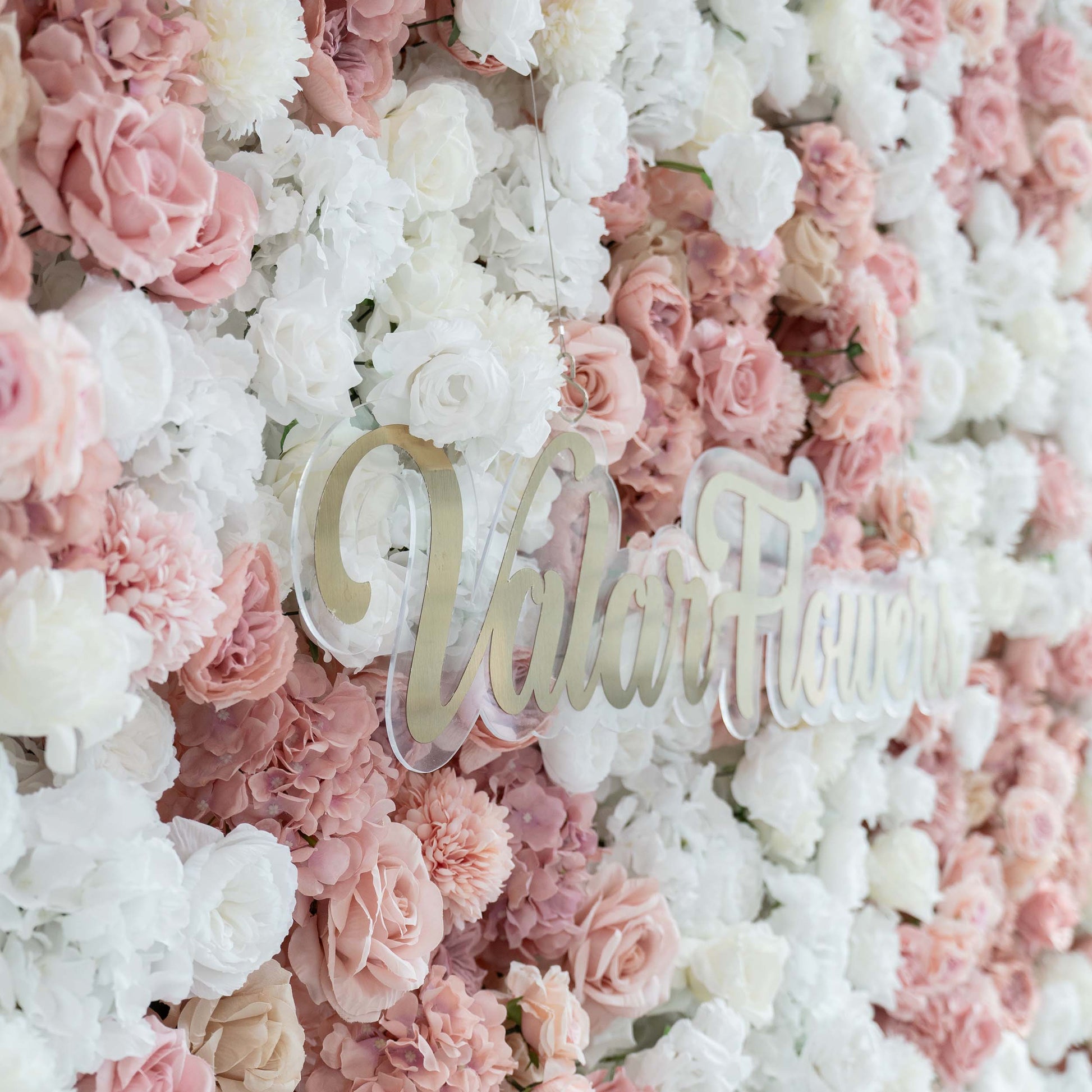 A close - up of a floral wall by ValarFlowers showcases a blend of white and pink artificial roses and hydrangeas, with the brand's name prominently displayed in gold. 