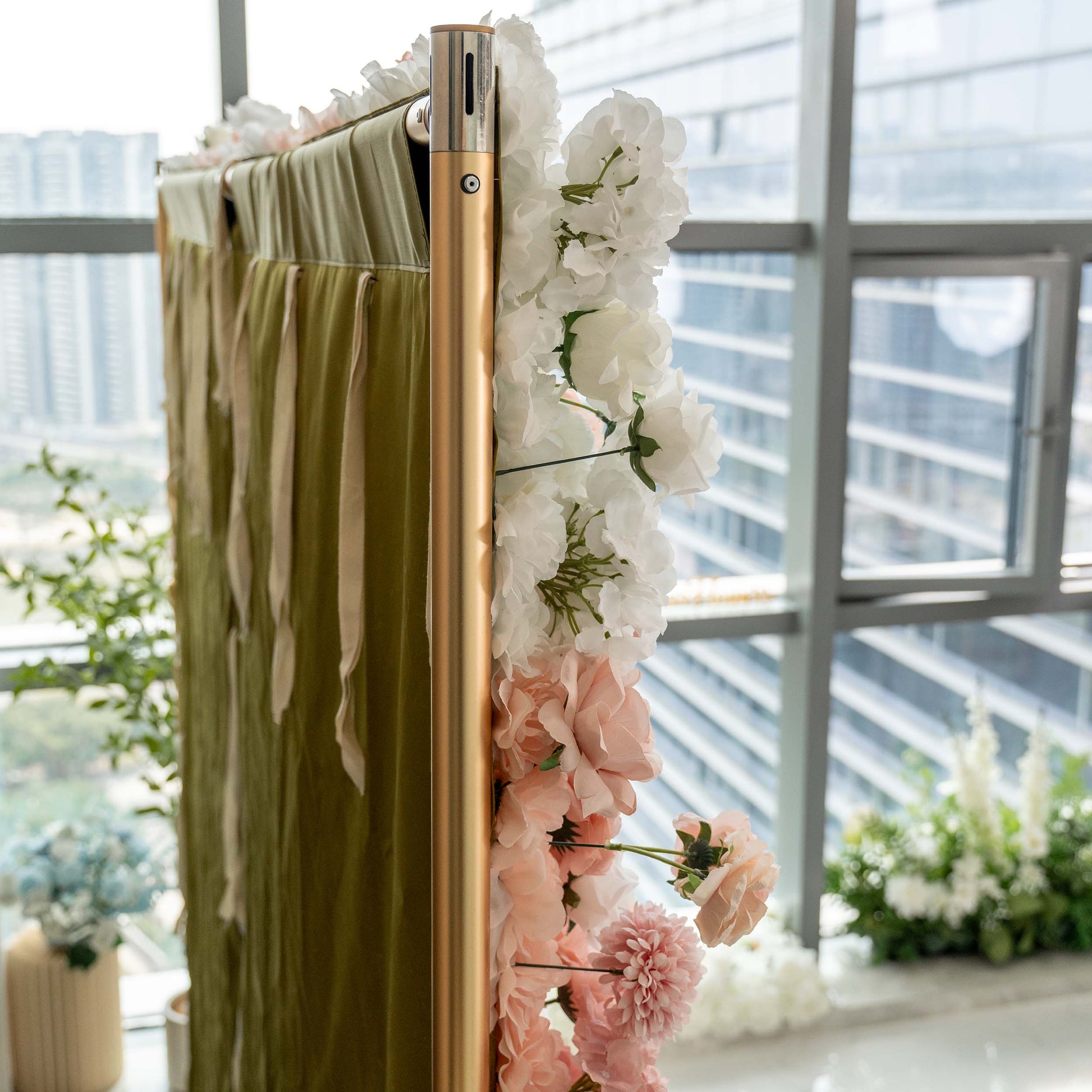 A close - up of a floral - adorned structure with a metallic pole. White and pink artificial flowers cascade over a green fabric panel, set against a window with a city view. 