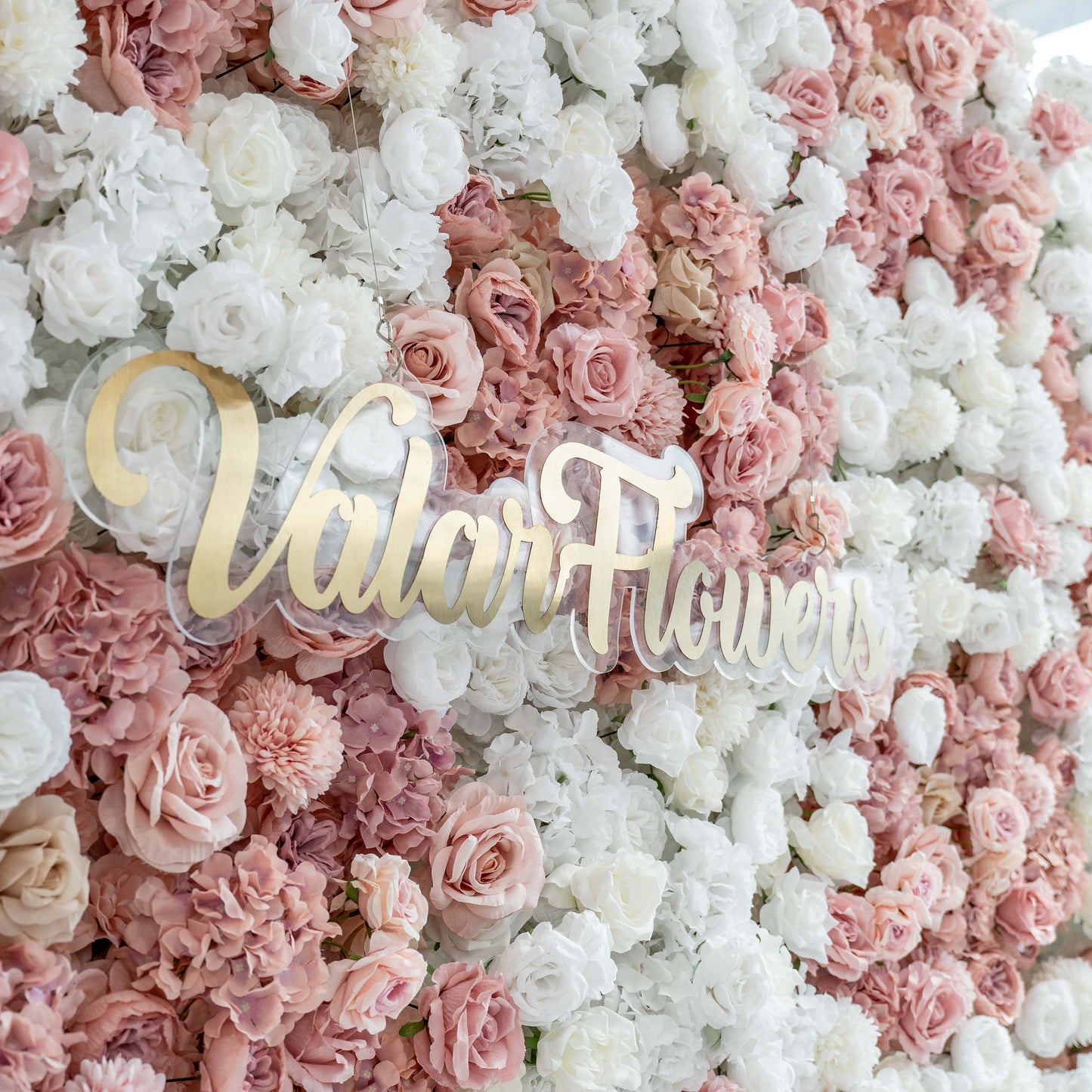 ValarFlowers' floral wall showcases a lush arrangement of white and pastel - pink roses and hydrangeas, with the brand name prominently displayed in gold. 
