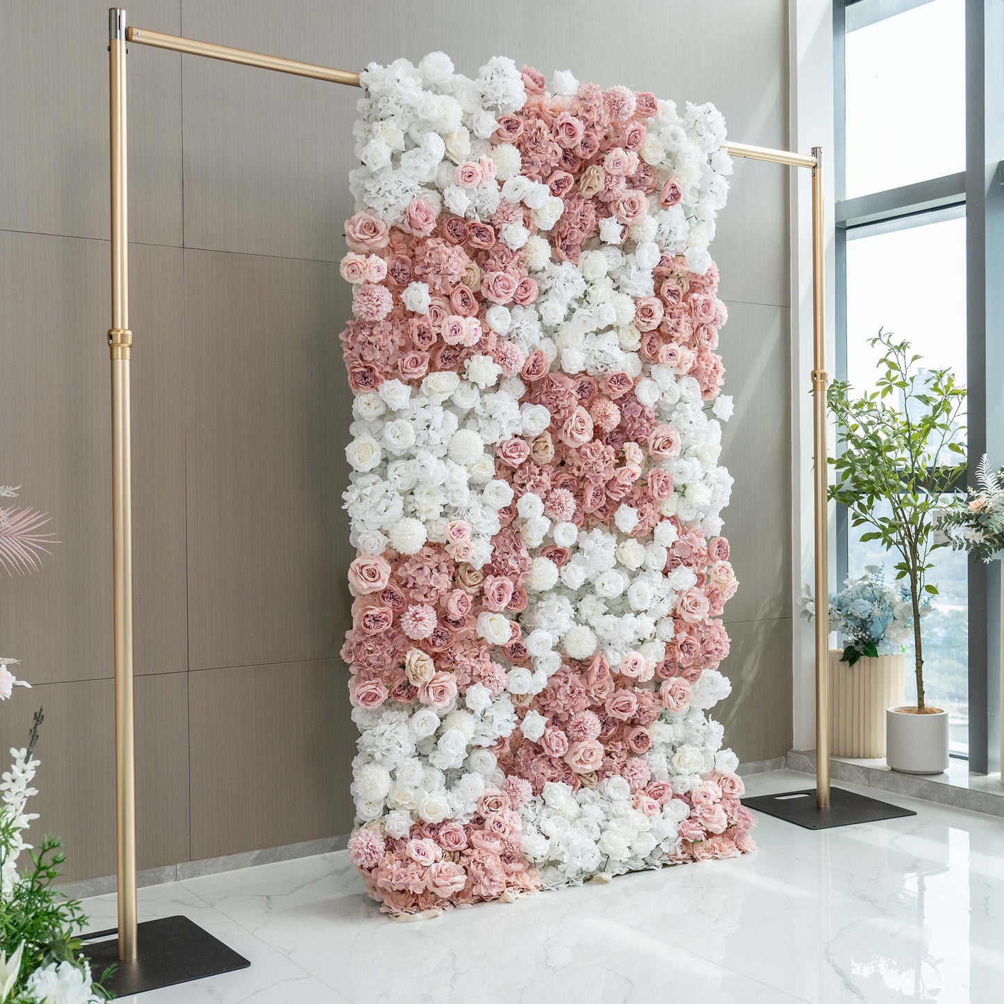A vertical floral wall with an artful mix of white and pink artificial roses and hydrangeas, framed by gold - toned poles, set in a well - lit room with a window view. 