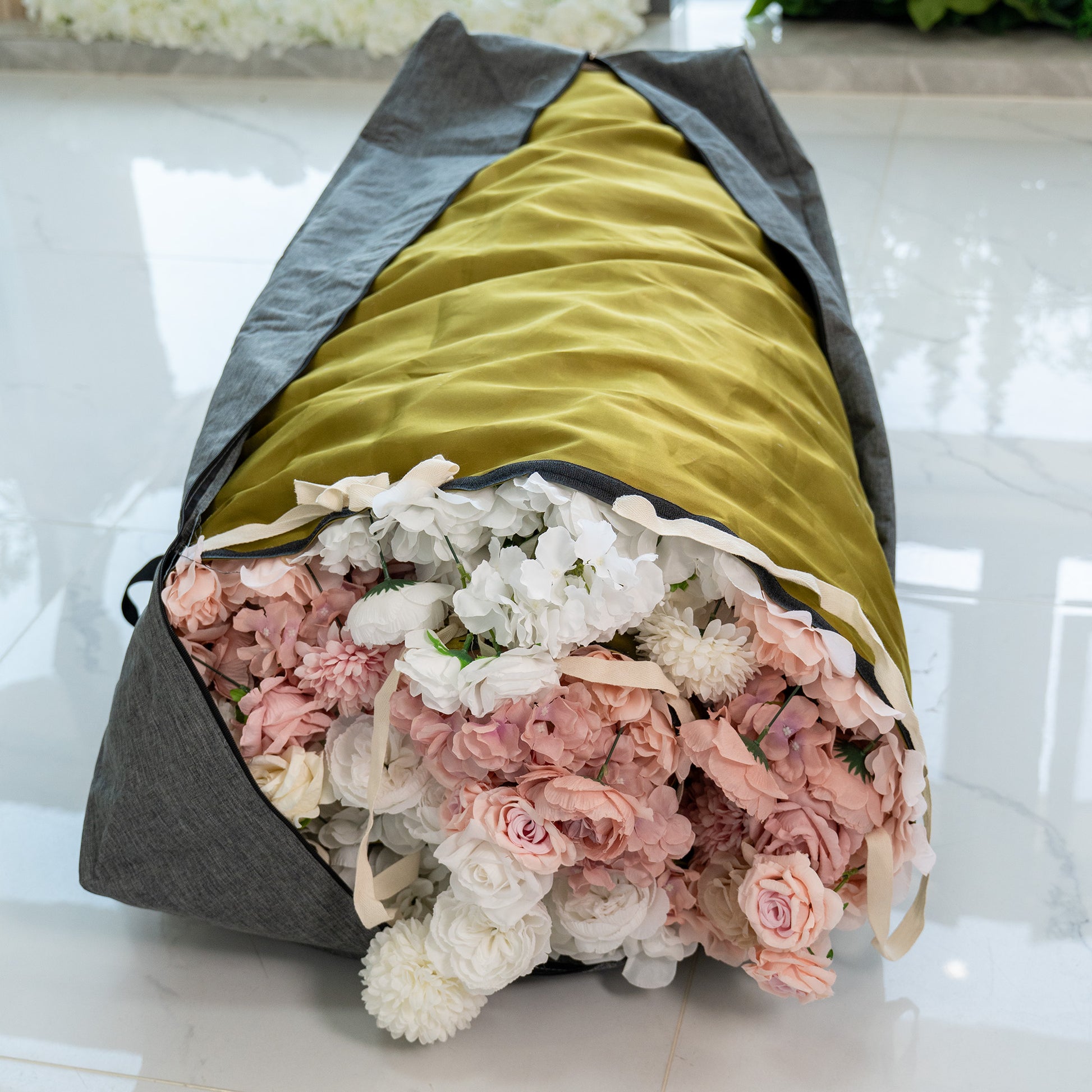 This image shows a storage bag designed for flower walls. The bag has a gray exterior with a mustard - colored interior. It is partially open, revealing a generous collection of artificial flowers in soft hues of pink, white, and light purple. The flowers appear to be well - packed inside. The bag also has straps, indicating it is portable, making it convenient for storing and transporting flower wall decorations. 