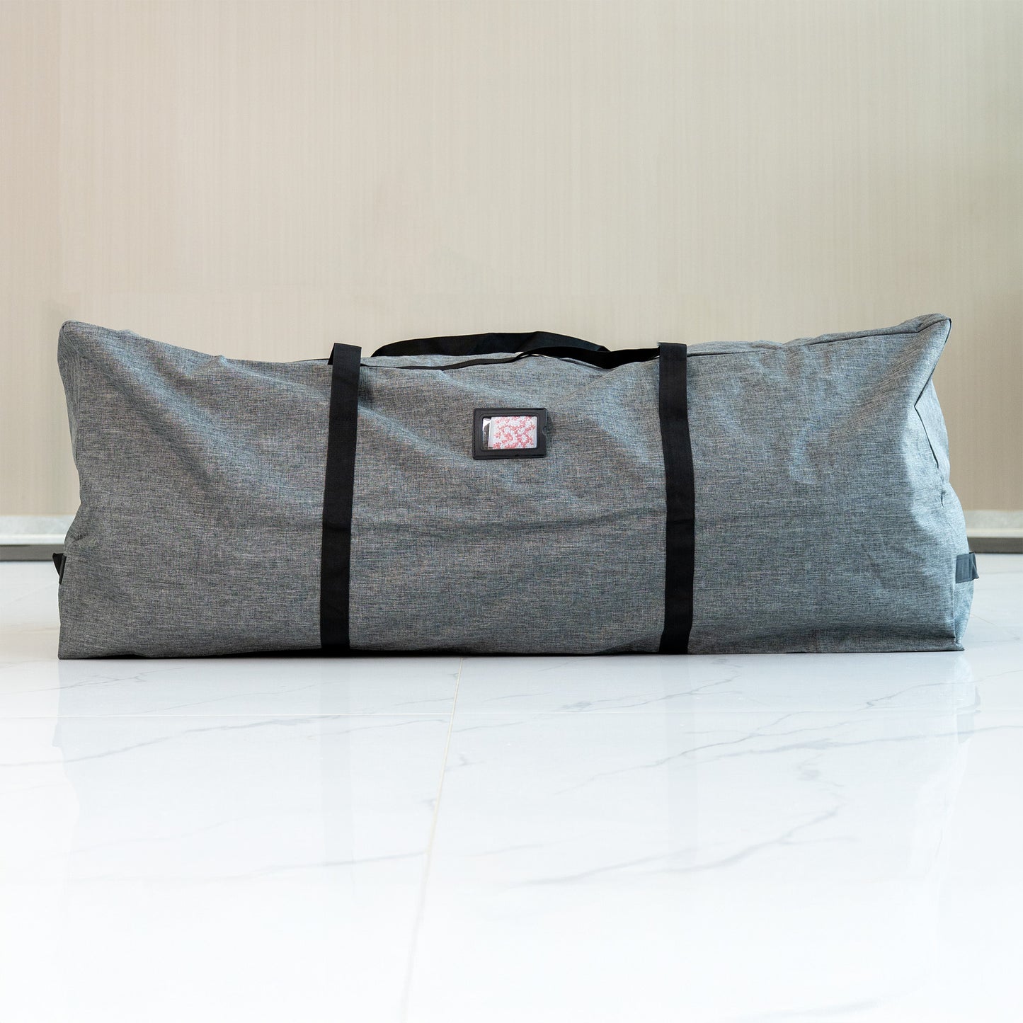 This is a storage bag for flower walls. It is large and rectangular, with a heathered gray fabric exterior. The bag is equipped with sturdy black carrying straps and features a small rectangular label patch with a pink - patterned insert. It is placed on a white marble - tiled floor against a neutral - colored wall, suggesting a clean and modern setting. The bag is designed for convenient storage and transport of flower wall decorations. 