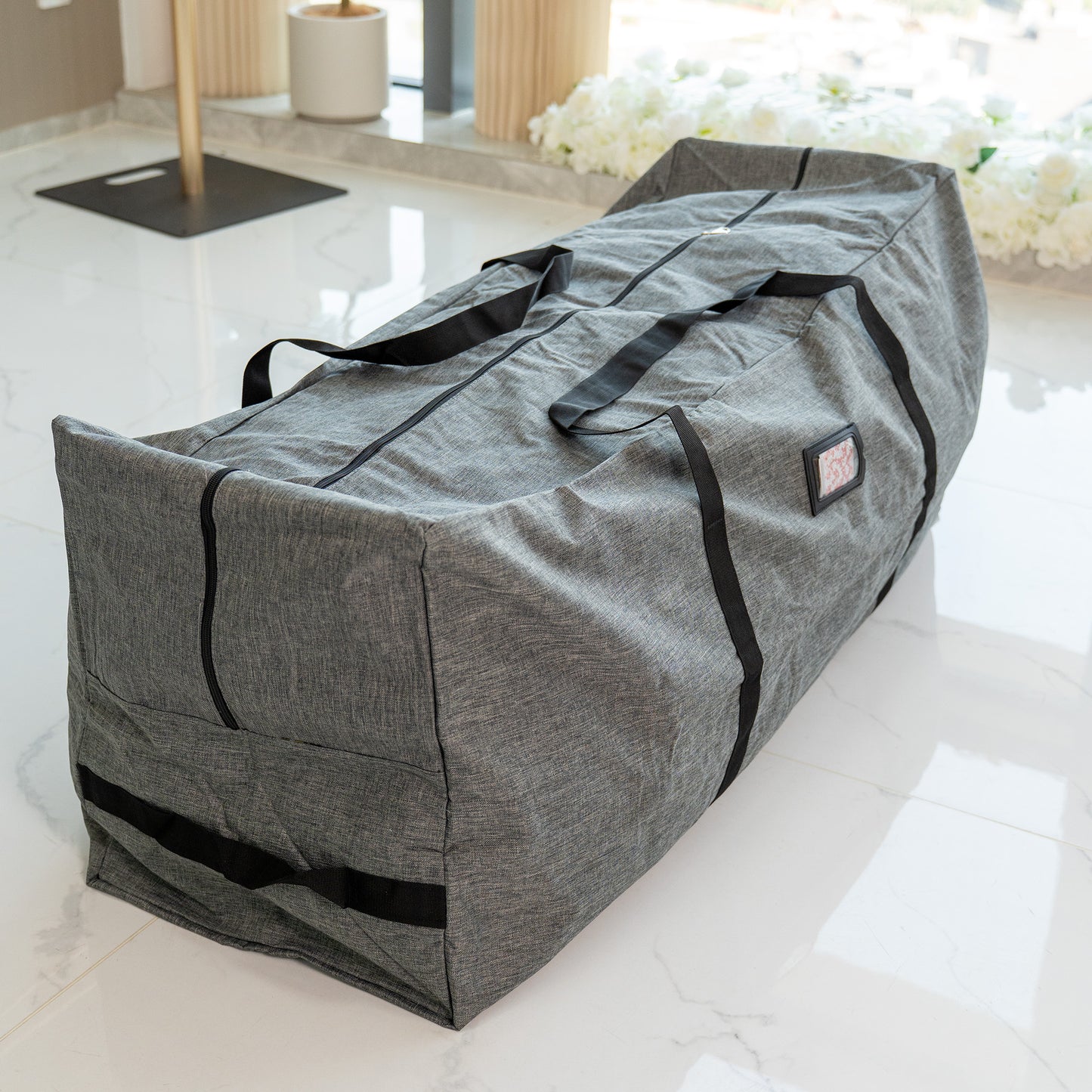 This image features a large, rectangular storage bag in a heathered gray color. The bag is equipped with sturdy black carrying straps and has a zipper running along its length, suggesting it can be opened wide for easy access. There is a small, rectangular label patch on the side of the bag. The bag is placed on a sleek, white marble - tiled floor, with a background that includes some white floral arrangements and modern, light - colored furniture elements, giving it a clean and contemporary setting. 