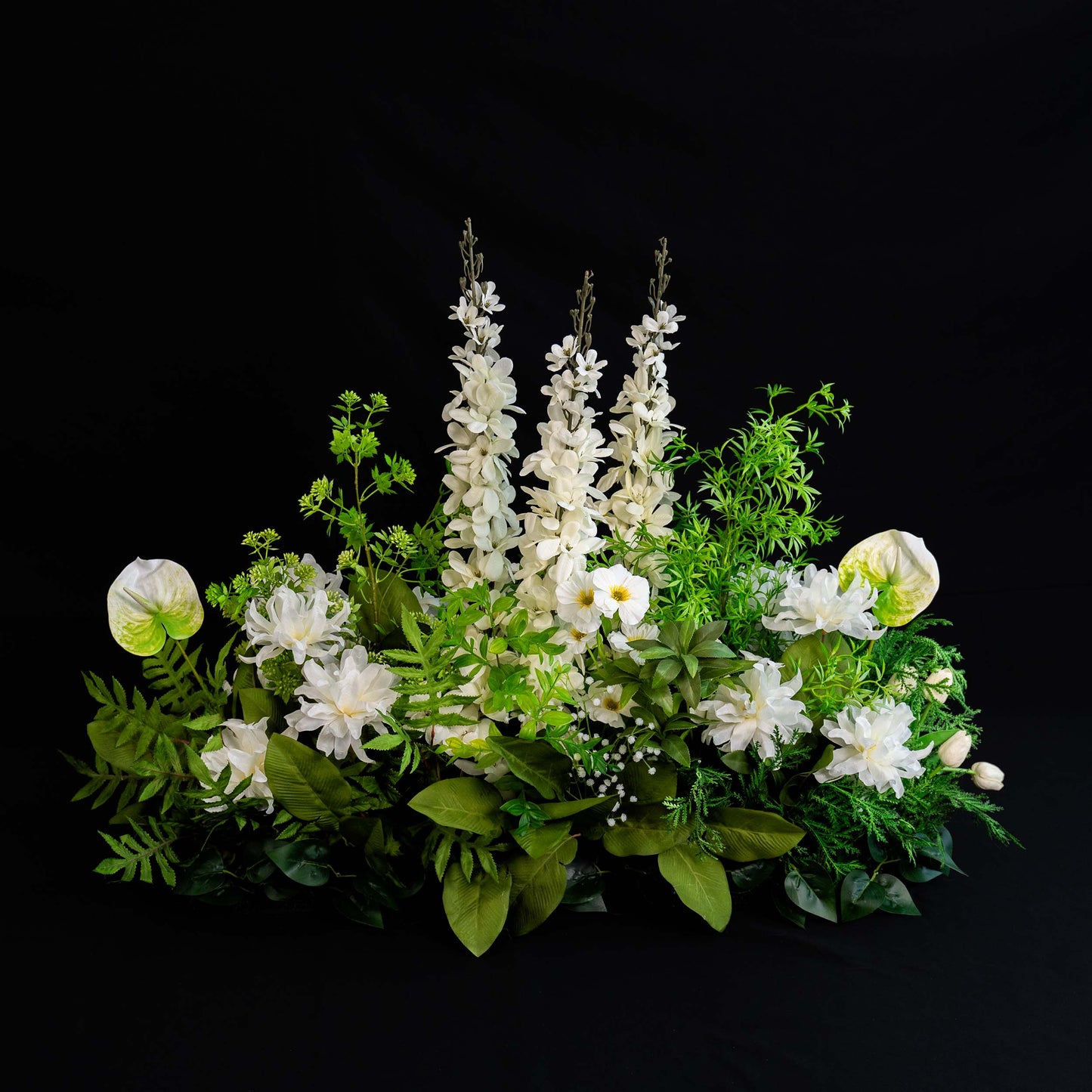 A floral arrangement features tall white blooms amidst an array of white flowers and lush green foliage, set against a stark black background for a striking contrast. 
