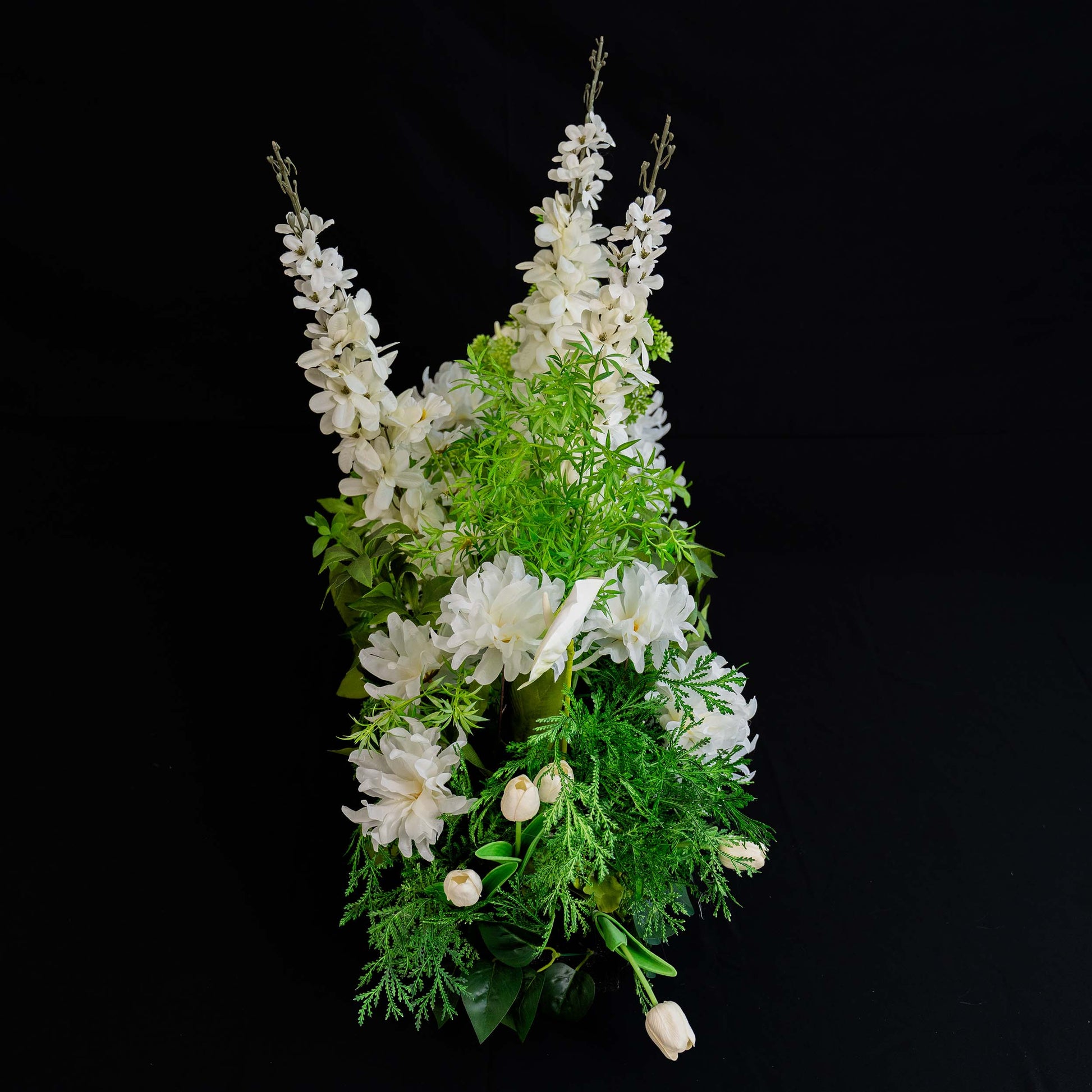 A side - view of a flower arrangement showcases tall white spire - like blooms, accompanied by large white flowers, white tulips, and lush green foliage against a black background. 
