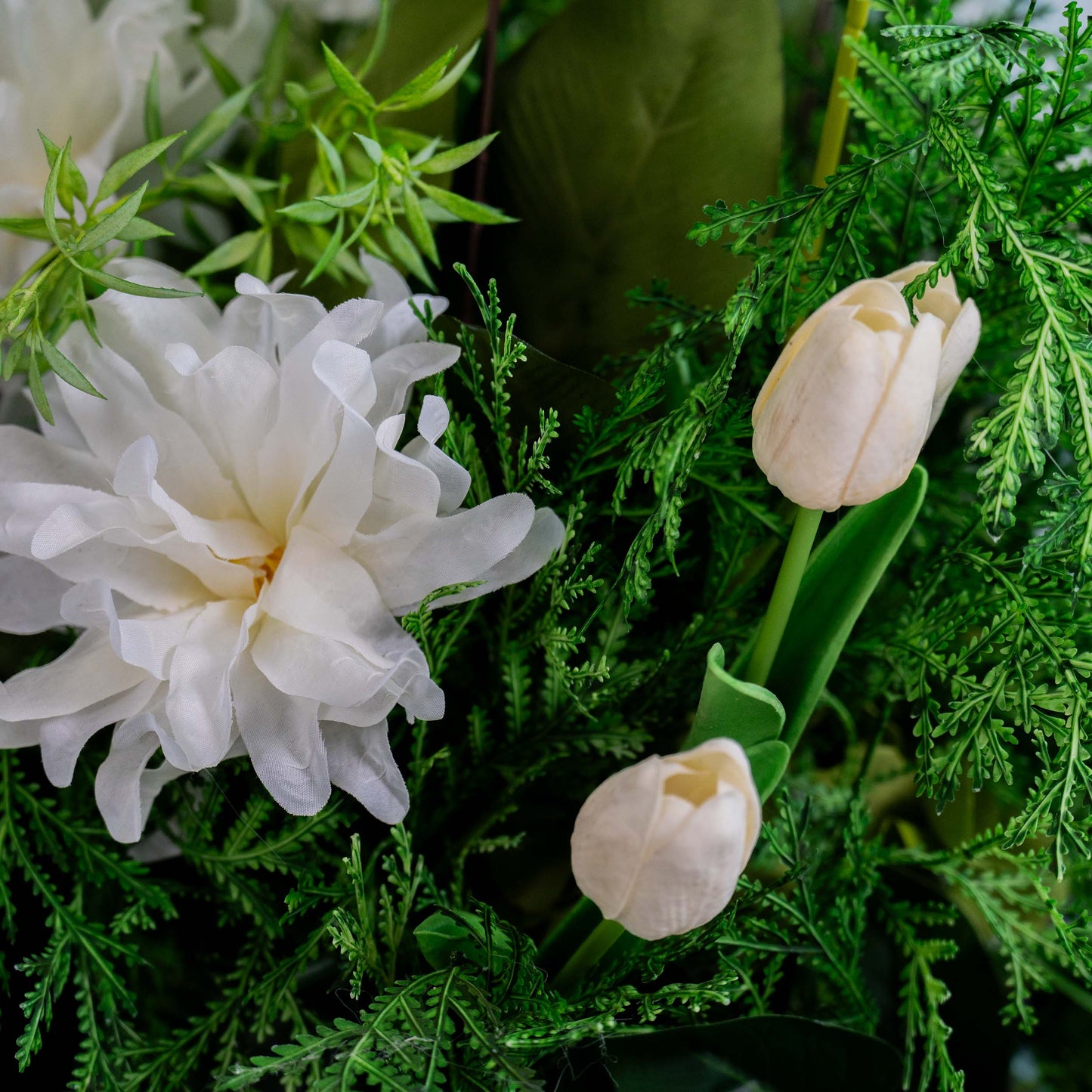 A close - up of a floral arrangement shows a large white bloom alongside two white tulips, all surrounded by lush green foliage, creating a fresh and delicate visual. 