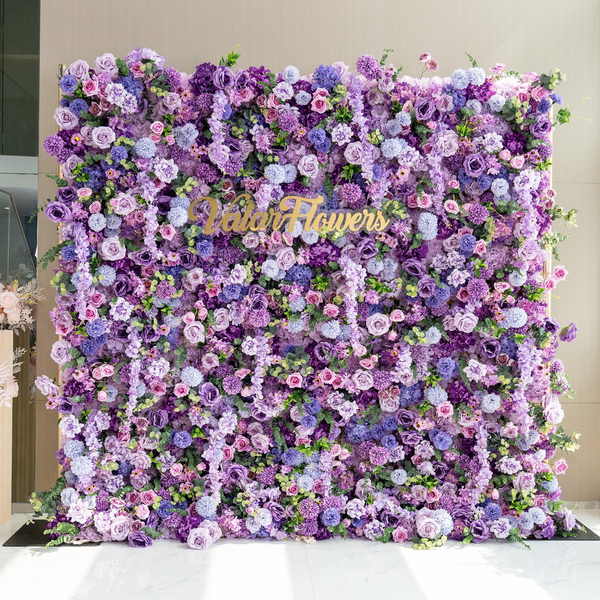 This image showcases a lush floral wall created by ValarFlowers. The wall is densely packed with an array of artificial flowers in various shades of purple, from deep violet to light lavender, along with hints of pink and green. Prominent flowers include roses, hydrangeas, and smaller bud - like blooms. The overall style is opulent and romantic, making it ideal for events like weddings or photo backdrops. 