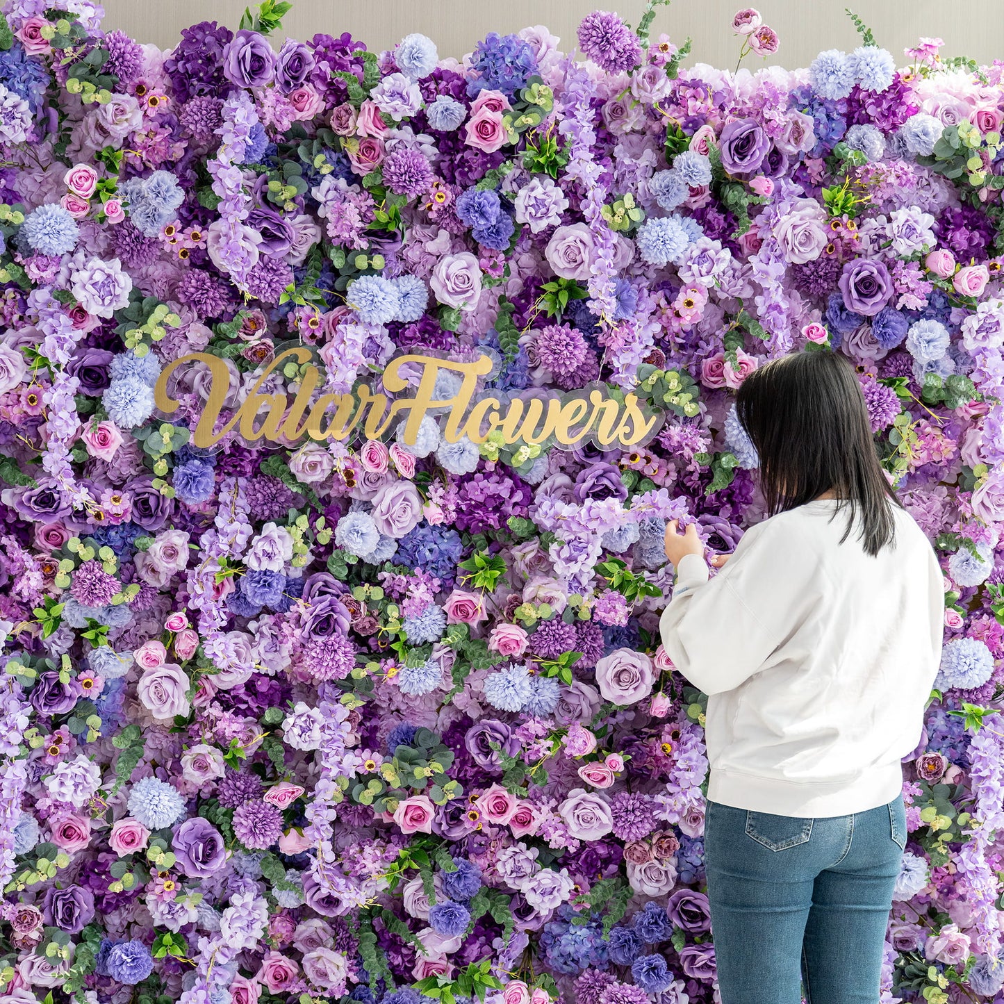 This image features a woman standing in front of a lush floral wall created by ValarFlowers. The wall is densely packed with an array of artificial flowers in various shades of purple, from deep violet to light lavender, along with hints of pink and green. Prominent flowers include roses, hydrangeas, and chrysanthemum - like blooms. The overall style is rich and romantic, perfect for events or photo opportunities. 