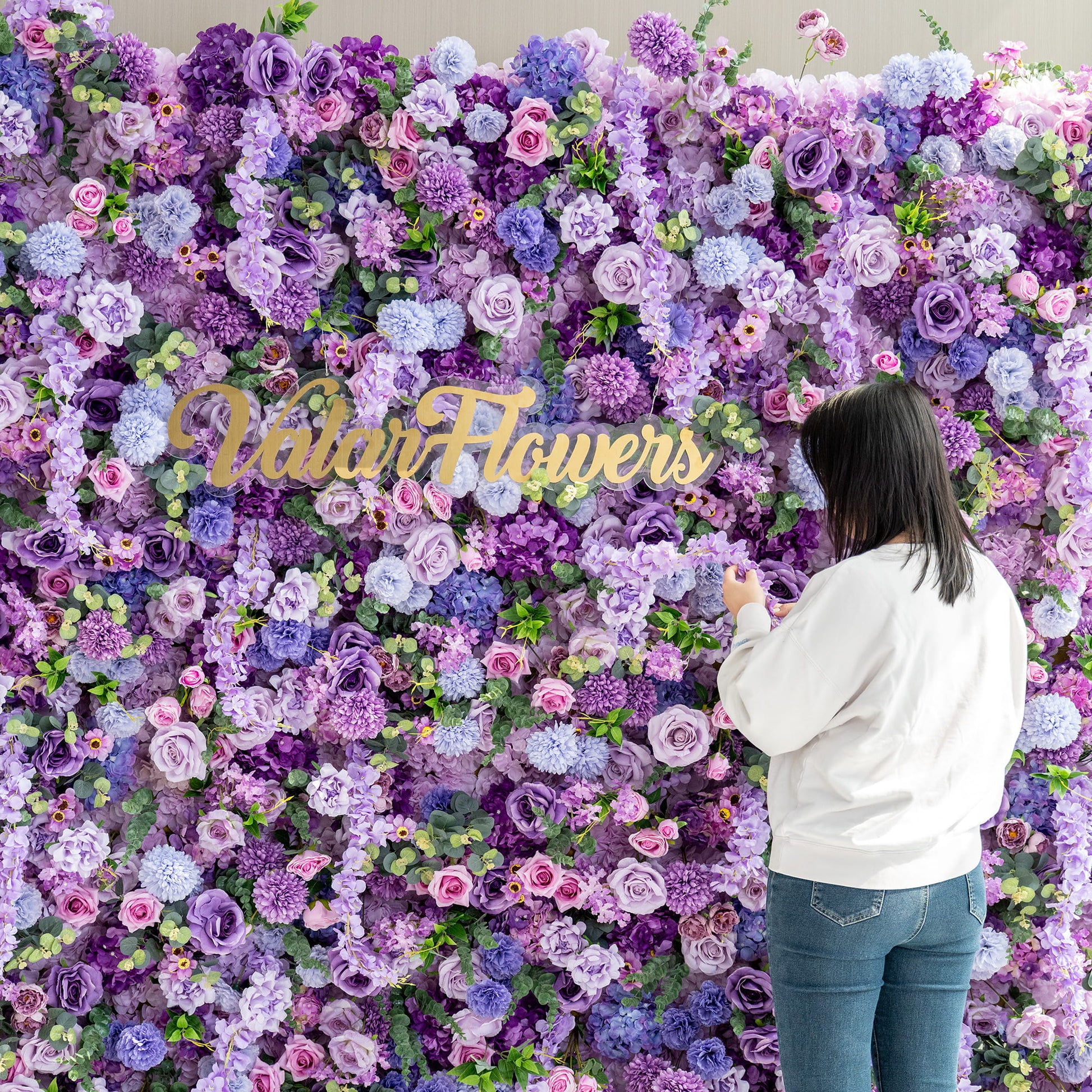 This image features a woman standing in front of a lush floral wall created by ValarFlowers. The wall is densely packed with an array of artificial flowers in various shades of purple, from deep violet to light lavender, along with hints of pink and green. Prominent flowers include roses, hydrangeas, and chrysanthemum - like blooms. The overall style is rich and romantic, perfect for events or photo opportunities. 
