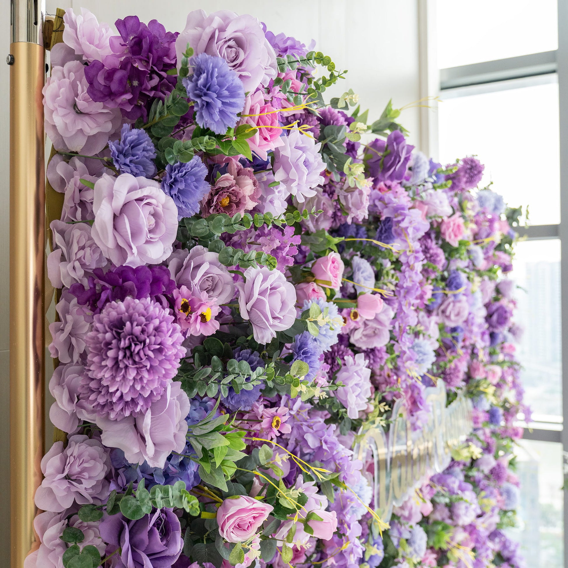 This image showcases a lush floral arrangement on a wall. It features an array of artificial flowers in various shades of purple, from deep violet to light lavender, along with hints of pink. Prominent flowers include roses, large - petaled blooms resembling peonies or chrysanthemums, and clusters of smaller flowers. Interspersed greenery adds a fresh touch, creating a rich, romantic, and visually appealing display. 