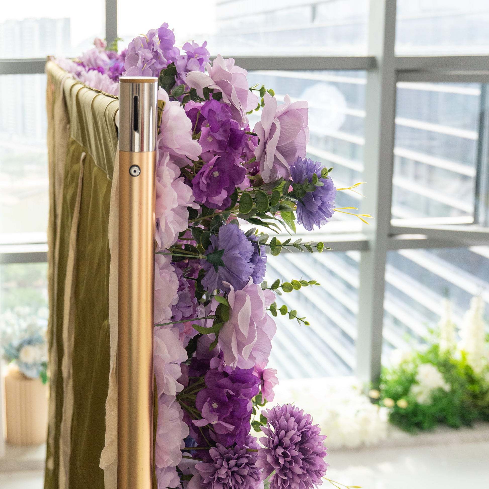 This image shows a close - up of a decorative setup featuring a golden metal pole, likely part of a frame or stand. Adjacent to the pole is a lush arrangement of artificial flowers in various shades of purple, from light lavender to deep violet. The flowers are interspersed with green foliage, creating a rich and vibrant display. In the background, there is a glimpse of a modern building through large windows, suggesting an indoor, contemporary setting. 