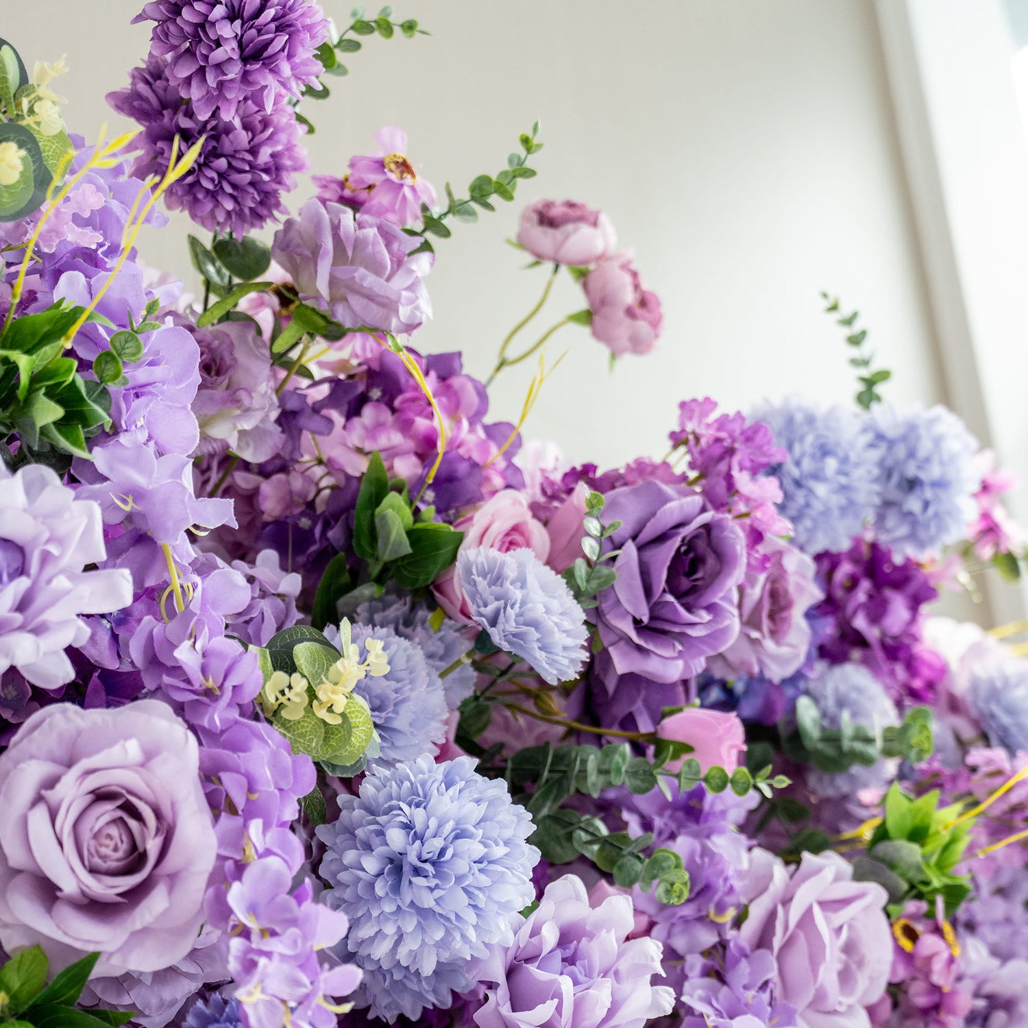 This image showcases a lush arrangement of artificial flowers in various shades of purple, ranging from deep violet to light lavender, with hints of blue and pink. Prominent flowers include roses, hydrangeas, and chrysanthemum - like blooms. Interspersed greenery adds a fresh touch. The overall style is romantic and opulent, creating a visually appealing and dreamy floral display. 
