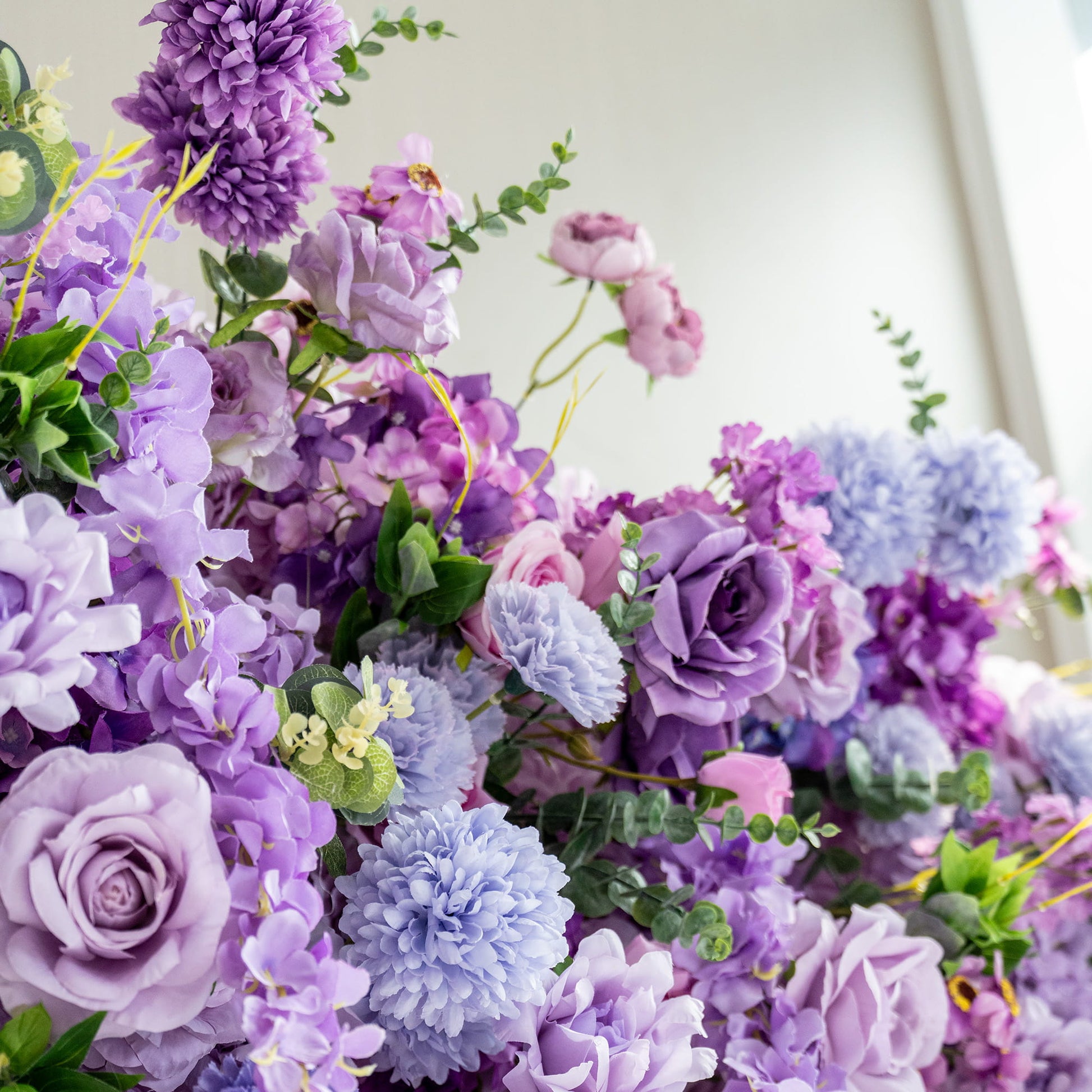 This image showcases a lush arrangement of artificial flowers in various shades of purple, ranging from deep violet to light lavender, with hints of blue and pink. Prominent flowers include roses, hydrangeas, and chrysanthemum - like blooms. Interspersed greenery adds a fresh touch. The overall style is romantic and opulent, creating a visually appealing and dreamy floral display. 