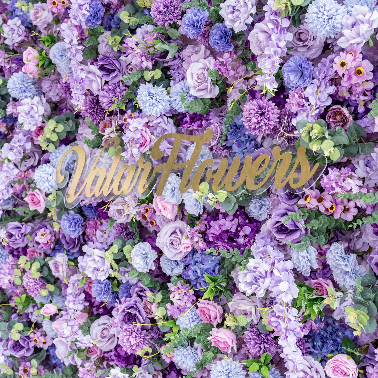 This image showcases a rich, textured floral wall with a captivating blend of artificial flowers in various shades of purple, from deep violet to light lavender, along with hints of pink and blue. Prominent flowers include roses, hydrangeas, and chrysanthemum - like blooms, interspersed with greenery. In the center, the golden text "ValarFlowers" adds an elegant touch, enhancing the overall romantic and opulent aesthetic. 