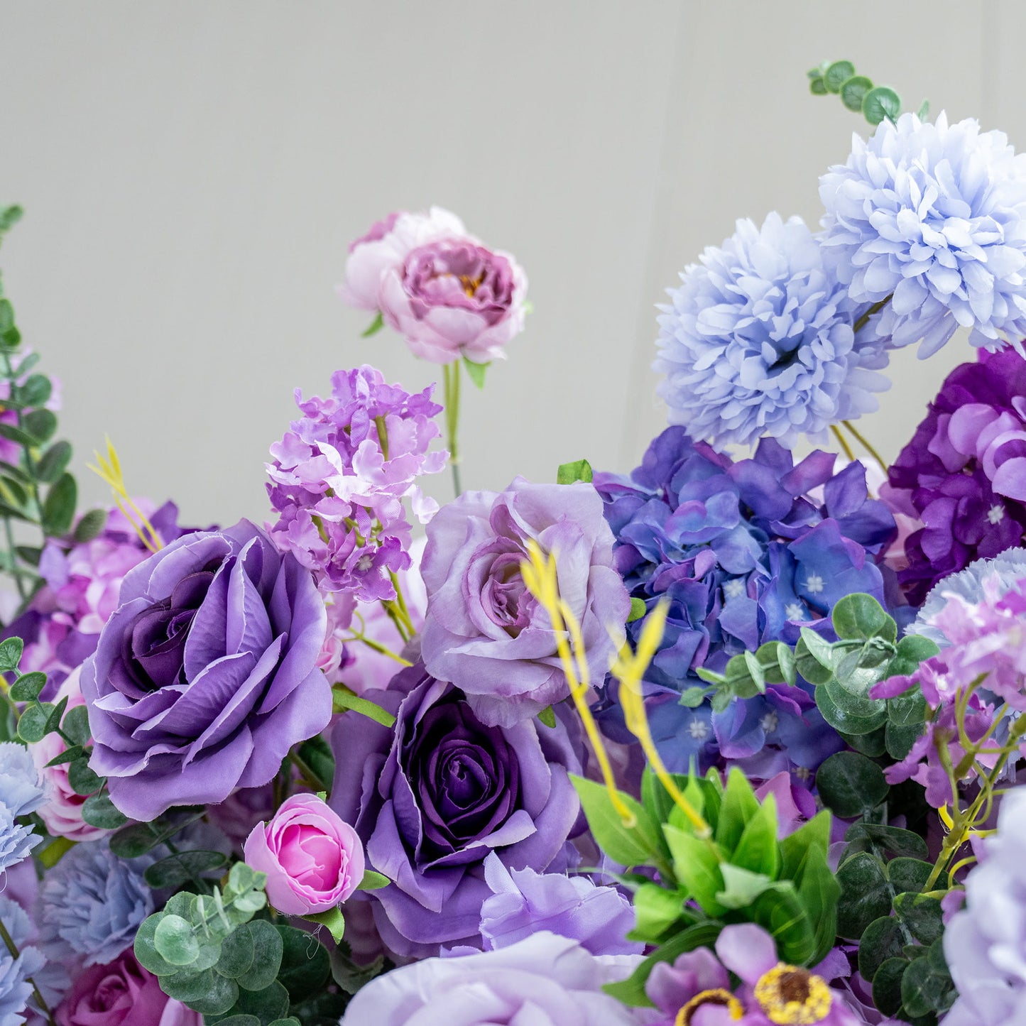 This image showcases a vibrant floral arrangement featuring an assortment of artificial flowers. Predominant are roses in various shades of purple, from deep violet to light lavender, accompanied by soft - pink roses. There are also large, fluffy hydrangeas in light blue and purple hues, along with clusters of lilac - colored flowers. The greenery adds a fresh touch, creating a romantic and elegant style. 
