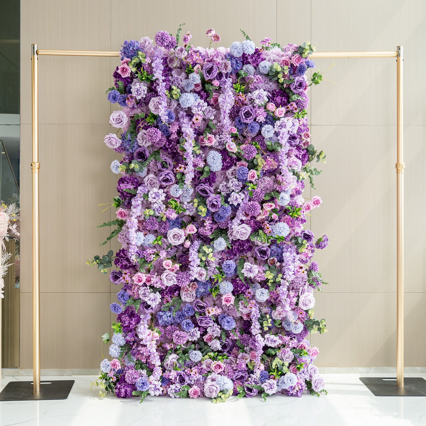 This image showcases a stunning 1.2 x 2.4m floral wall supported by a golden stand. The wall is densely adorned with an array of artificial flowers in various shades of purple, from deep violet to light lavender, along with hints of pink and green. Prominent flowers include roses, hydrangeas, and chrysanthemum - like blooms. The overall style is opulent and romantic, making it perfect for events or as a photo backdrop. 
