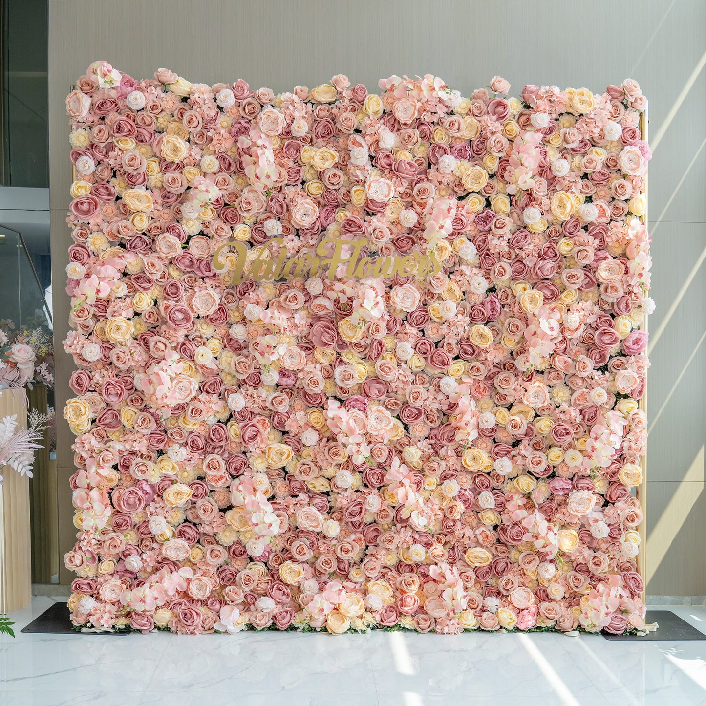 The image features a large flower wall backdrop filled with artificial roses in shades of pink, blush, and cream. In the center, the branding “Valarflowers” in gold - toned letters stands out. The style is opulent and romantic. 