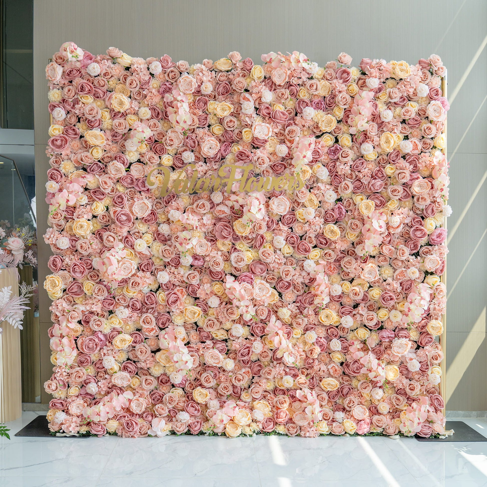 The image features a large flower wall backdrop filled with artificial roses in shades of pink, blush, and cream. In the center, the branding “Valarflowers” in gold - toned letters stands out. The style is opulent and romantic. 