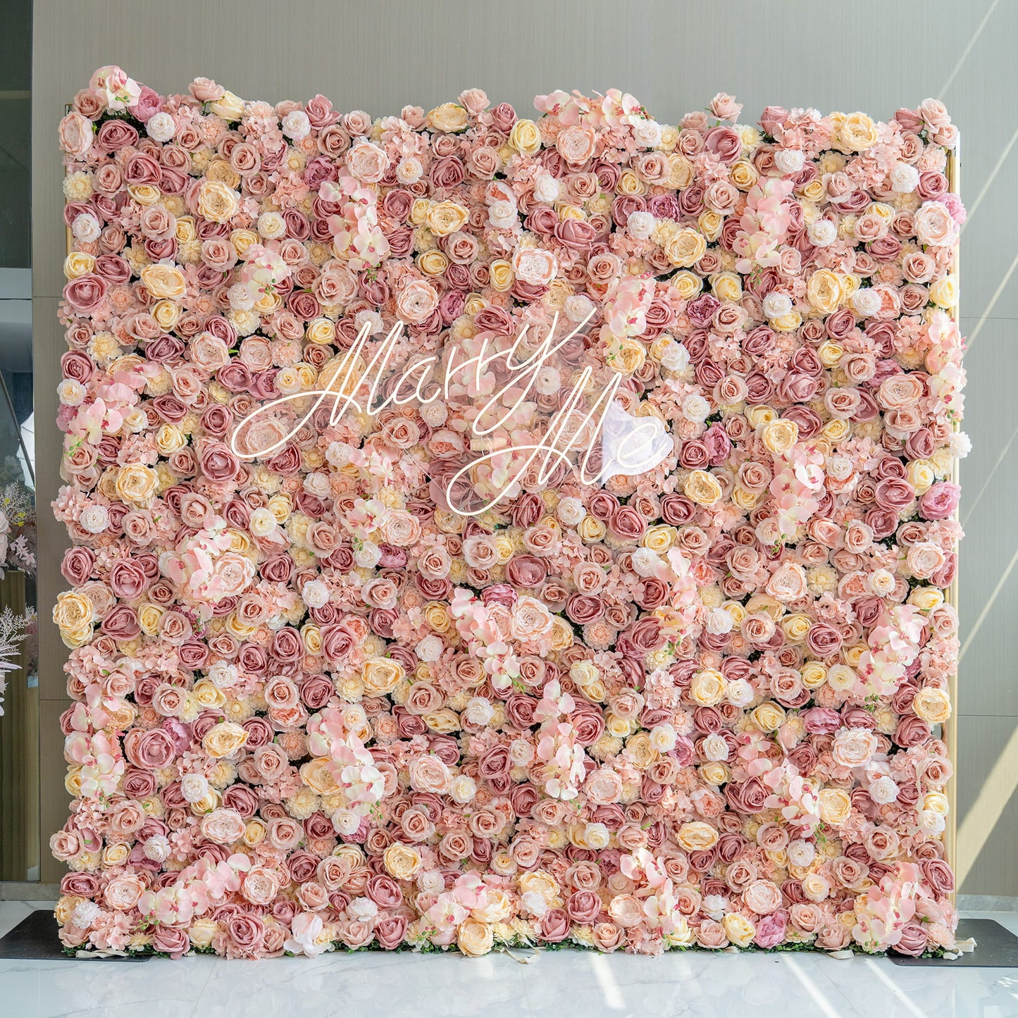 The image displays a large flower wall filled with artificial roses and some orchids in soft pink, peach, and cream hues. In the center, there's the text "Marry Me." The style is romantic and elegant. 