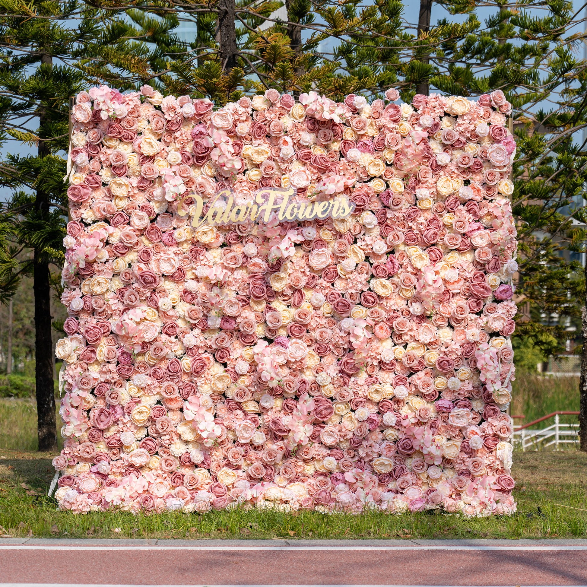 The image shows an outdoor flower wall mainly made of artificial roses in soft pink, peach, and cream colors. The “ValarFlowers” logo is in the middle. It has a romantic and elegant style. 