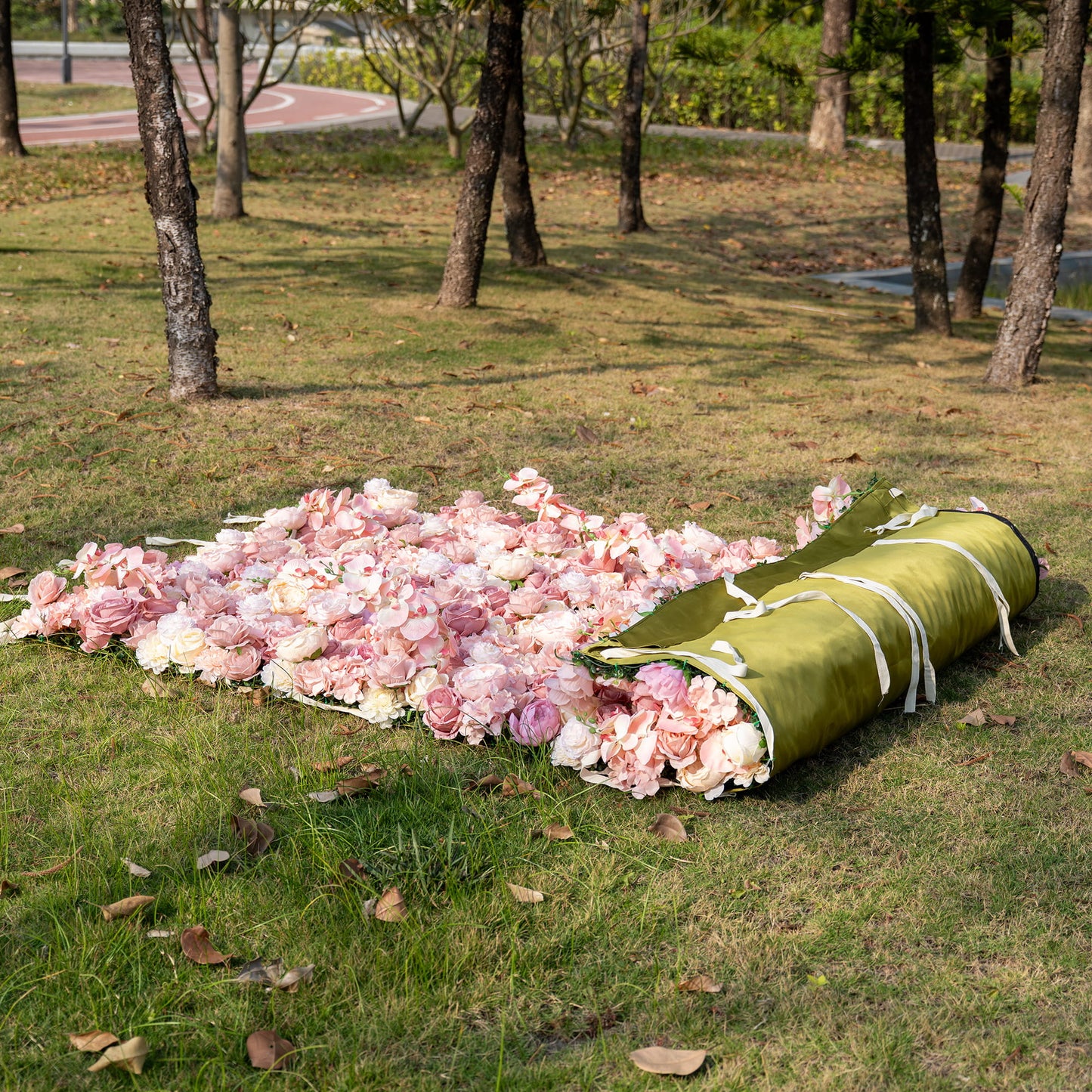 The image shows a rolled - up artificial flower panel on a grassy area outdoors. It's filled with roses and other blooms in soft pink and cream colors, giving a romantic and convenient decor style. 