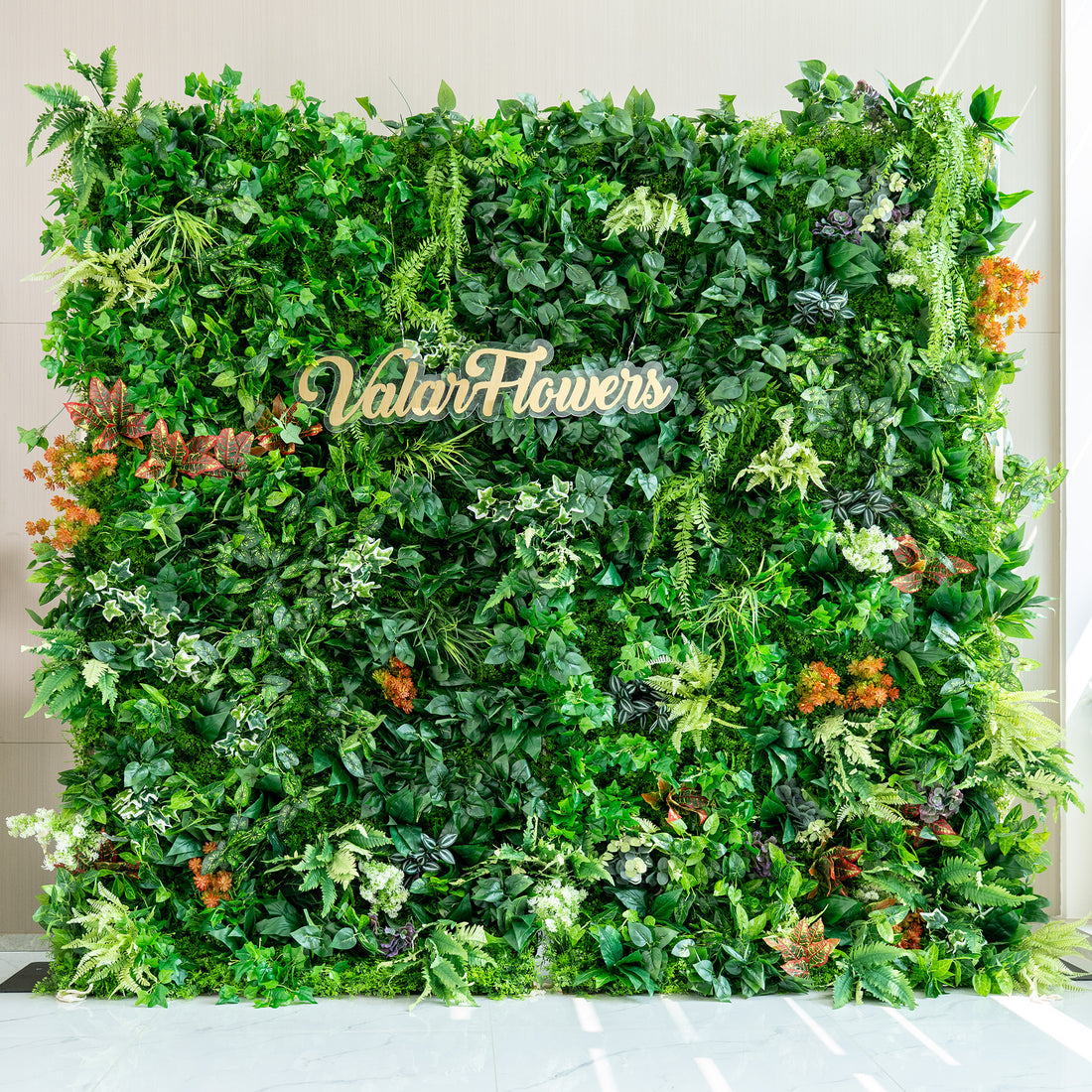 This image features a lush artificial green wall by ValarFlowers. The wall is densely packed with a variety of faux plants, including different shades of green foliage such as ivy - like leaves, ferns, and broad - leafed plants. There are also pops of color from orange and red - tinged plants, adding a vibrant contrast. The overall effect is a rich, natural - looking greenery backdrop, ideal for events or decor purposes. 