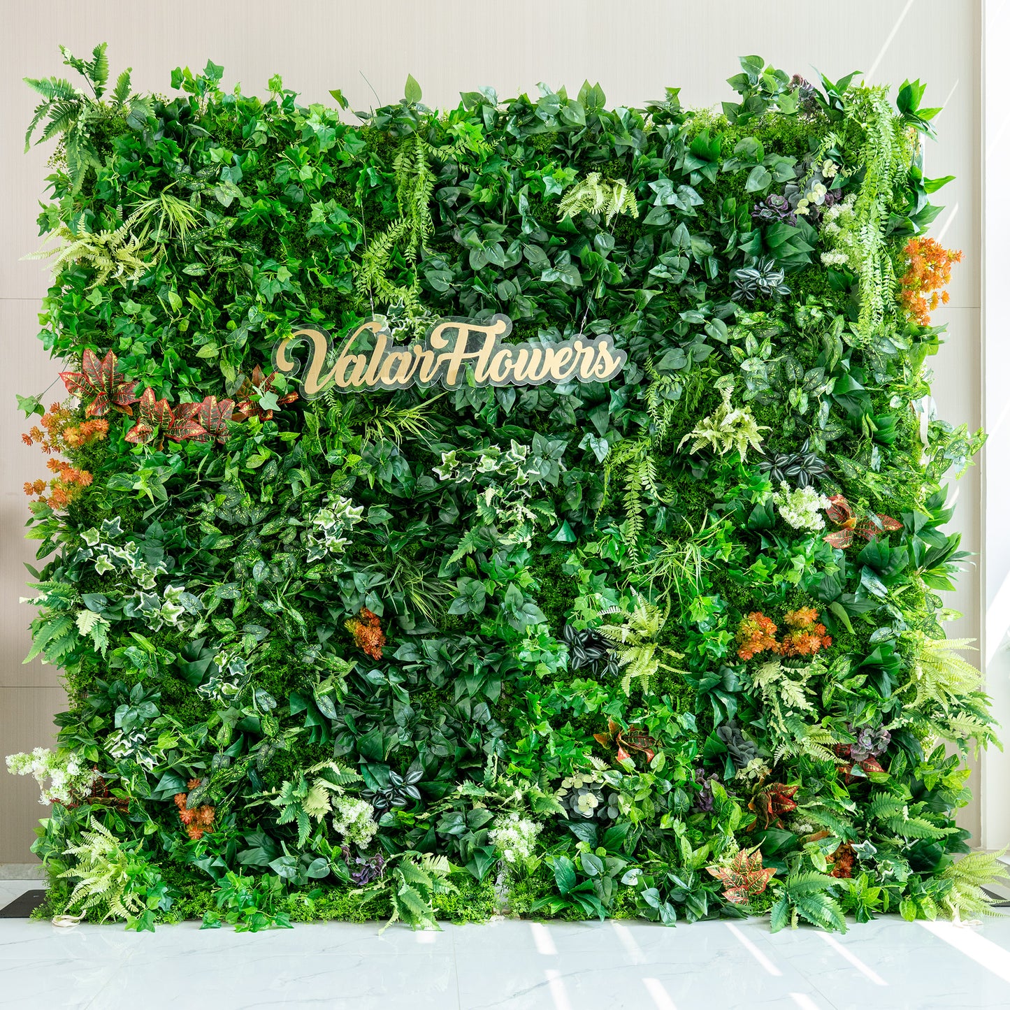 This image features a lush artificial green wall by ValarFlowers. The wall is densely packed with a variety of faux plants, including different shades of green foliage such as ivy - like leaves, ferns, and broad - leafed plants. There are also pops of color from orange and red - tinged plants, adding a vibrant contrast. The overall effect is a rich, natural - looking greenery backdrop, ideal for events or decor purposes. 