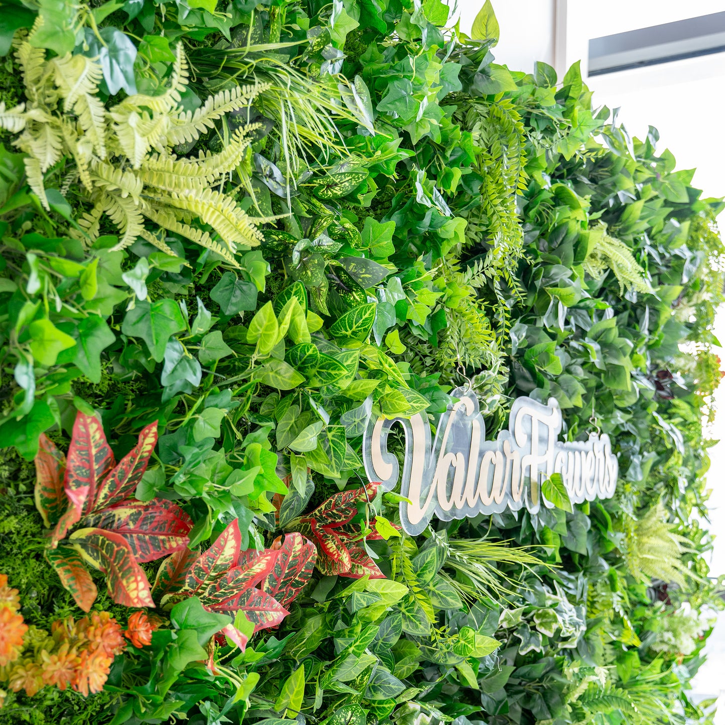 This image showcases a lush, vibrant green wall densely packed with a variety of artificial plants. The foliage includes different shades of green ferns, broad - leafed plants, and vines. Interspersed are striking red - and - green variegated leaves, adding splashes of color. The text "ValarFlowers" is prominently displayed, suggesting the creator or brand behind this greenery display. The overall effect is rich and natural - looking. 