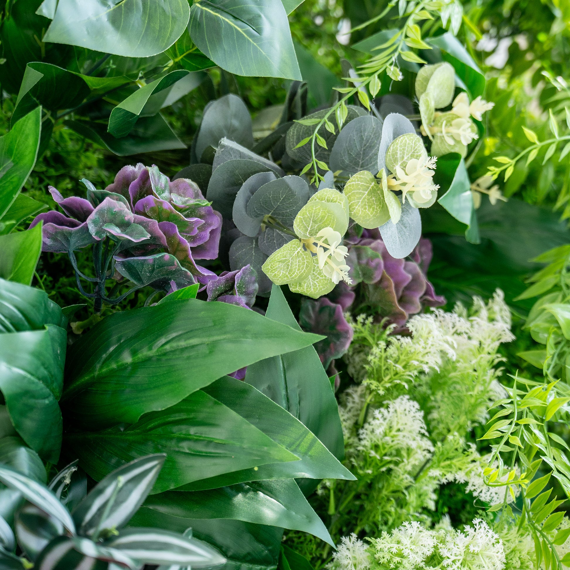 This image showcases a vibrant and lush arrangement of artificial plants. It features a mix of large green leaves, purple - hued foliage resembling hydrangeas, and clusters of white, delicate flowers. Interspersed are grey - green eucalyptus - like leaves with small white blossoms, adding texture and contrast. The overall scene is rich and lively, mimicking a natural garden setting. 