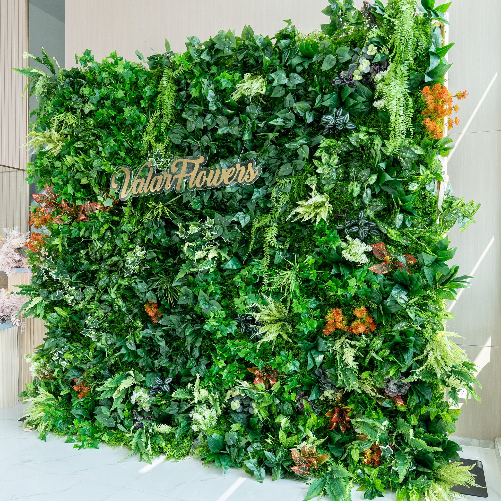 This image showcases a lush, vibrant green wall crafted by ValarFlowers. The wall is densely packed with a rich variety of artificial plants, featuring an array of green foliage in different shades and textures, from deep emerald to light lime. Interspersed are splashes of color from orange, red, and white - tinged plants, adding visual interest. The overall effect is a natural - looking, verdant backdrop, perfect for events or decor. 