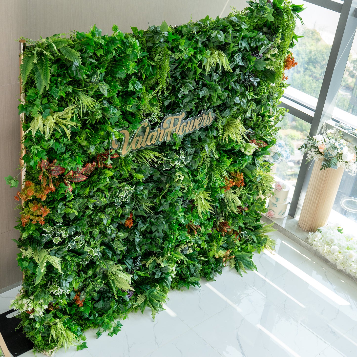 This image features a lush, artificial green wall by ValarFlowers. The wall is densely packed with a variety of faux plants, including different shades of green foliage such as ivy - like leaves, ferns, and broad - leafed plants. There are also splashes of color from orange and red - tinged plants, adding a vibrant contrast. The backdrop is set indoors near a window, with some floral arrangements visible nearby, enhancing the fresh and natural ambiance. 