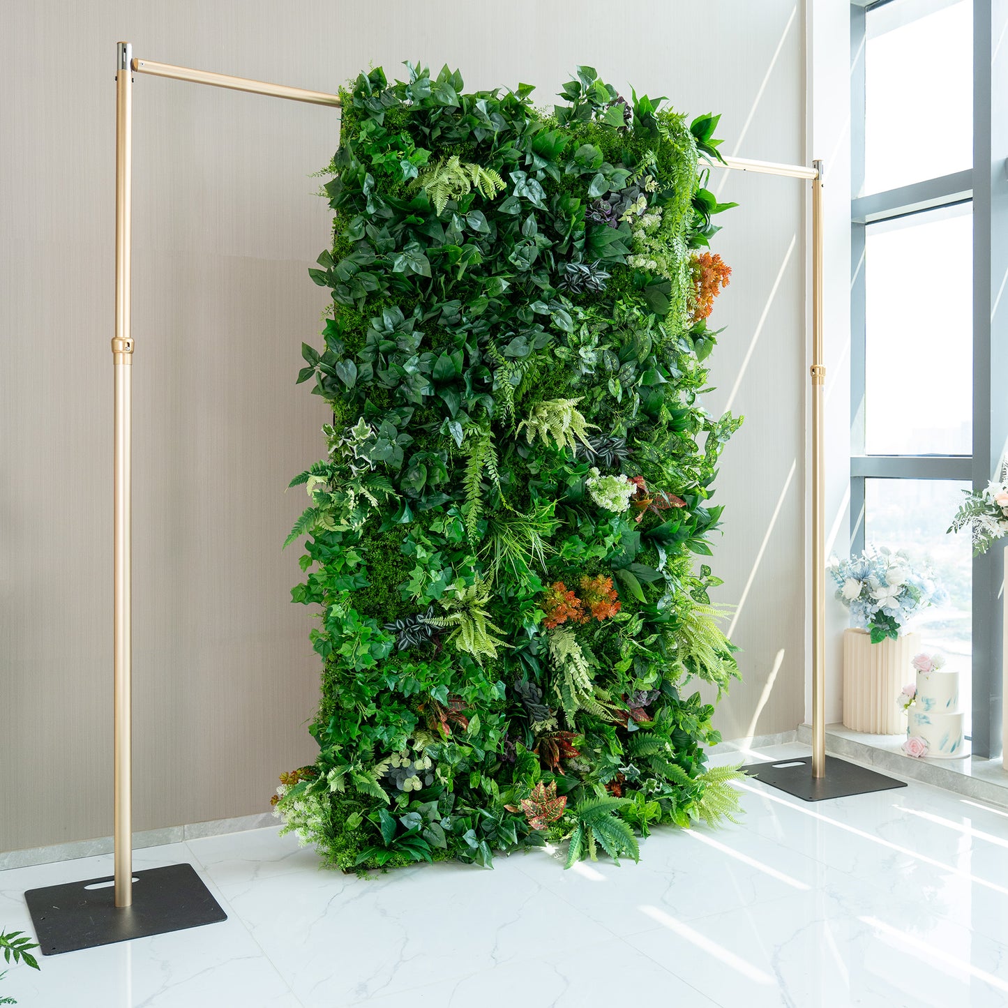 This image depicts a lush, artificial green wall supported by a metallic gold - colored frame. The wall is densely packed with a variety of faux plants, showcasing different shades of green foliage, from light lime to deep emerald. Splashes of color are added by red, orange, and purple - hued plants. The setup is indoors, with a neutral - toned wall in the background and a glimpse of a window with a floral arrangement beside it. 