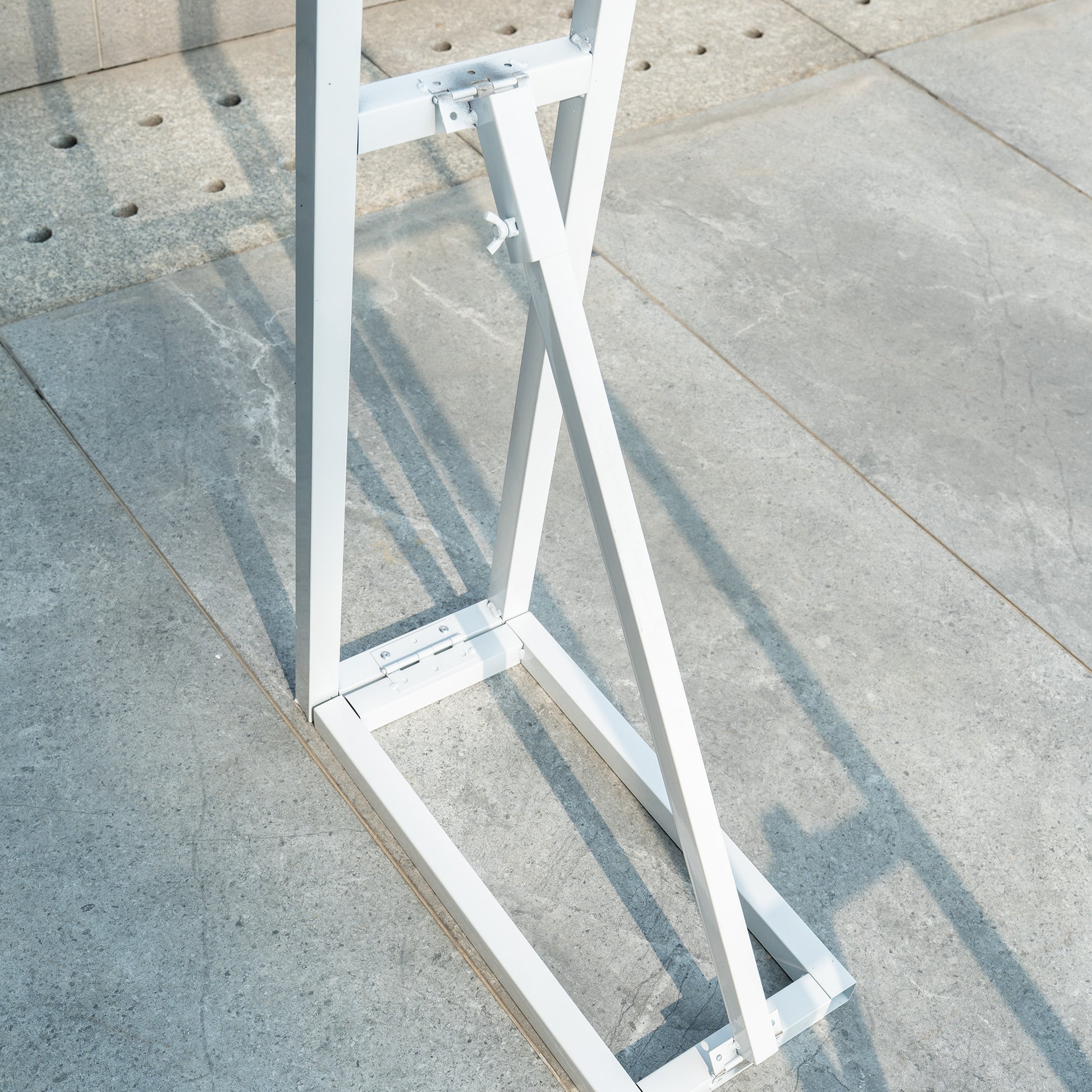 This backdrop stand is a metallic structure with a light - colored finish, likely made of aluminum or steel. It features a sturdy, geometric design with a rectangular base and diagonal support bars for stability. The stand has adjustable components, indicated by the bolts and hinges, allowing for easy setup and customization. It is placed on a concrete surface, suggesting it is designed for outdoor or event use. 
