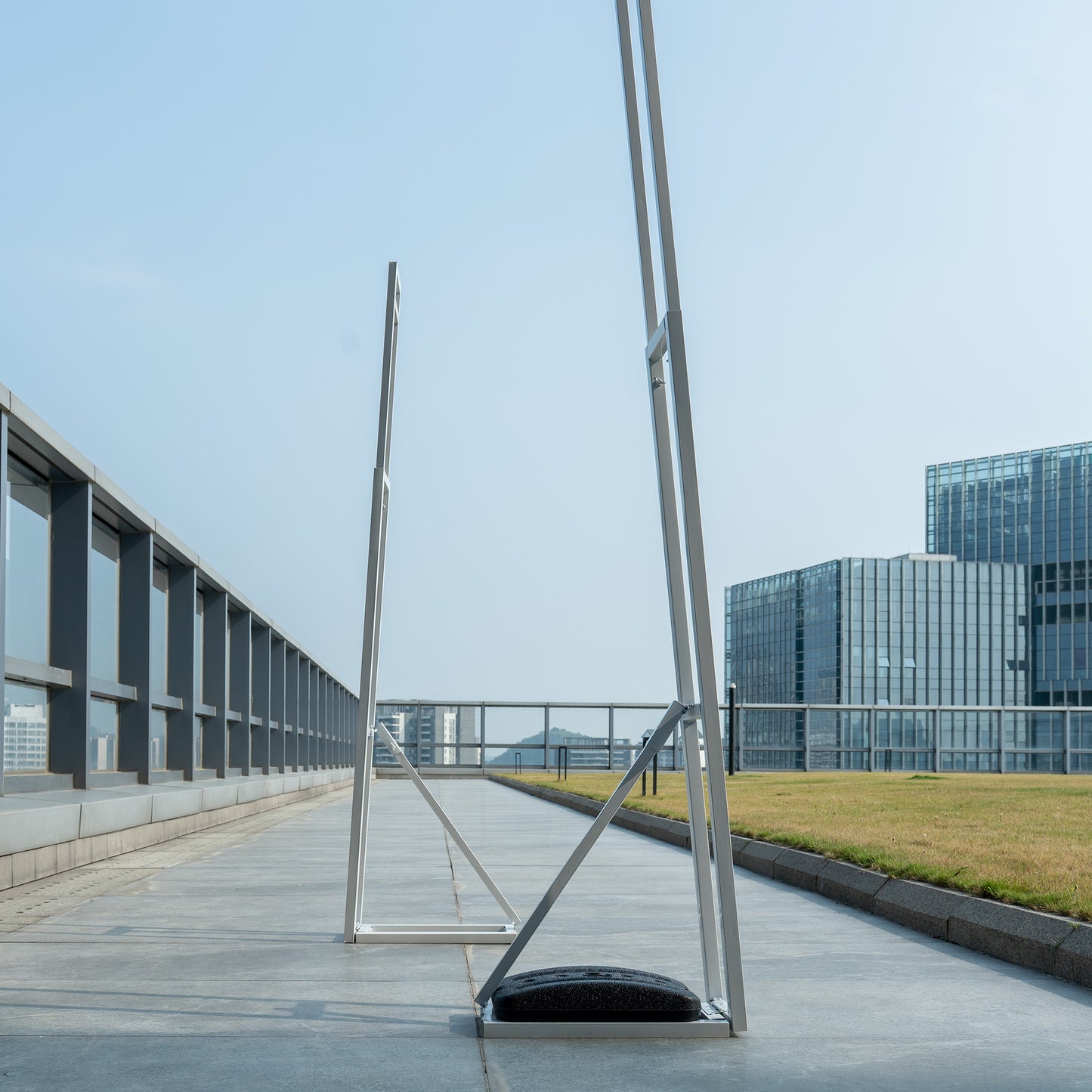 These backdrop stands are metallic structures positioned on an outdoor rooftop. They have a sleek, modern design with angled frames, giving them a contemporary aesthetic. The stands are equipped with black, weighted bases for stability, ensuring they can support floral or decorative backdrops for events. The urban cityscape in the background adds to the modern feel. 