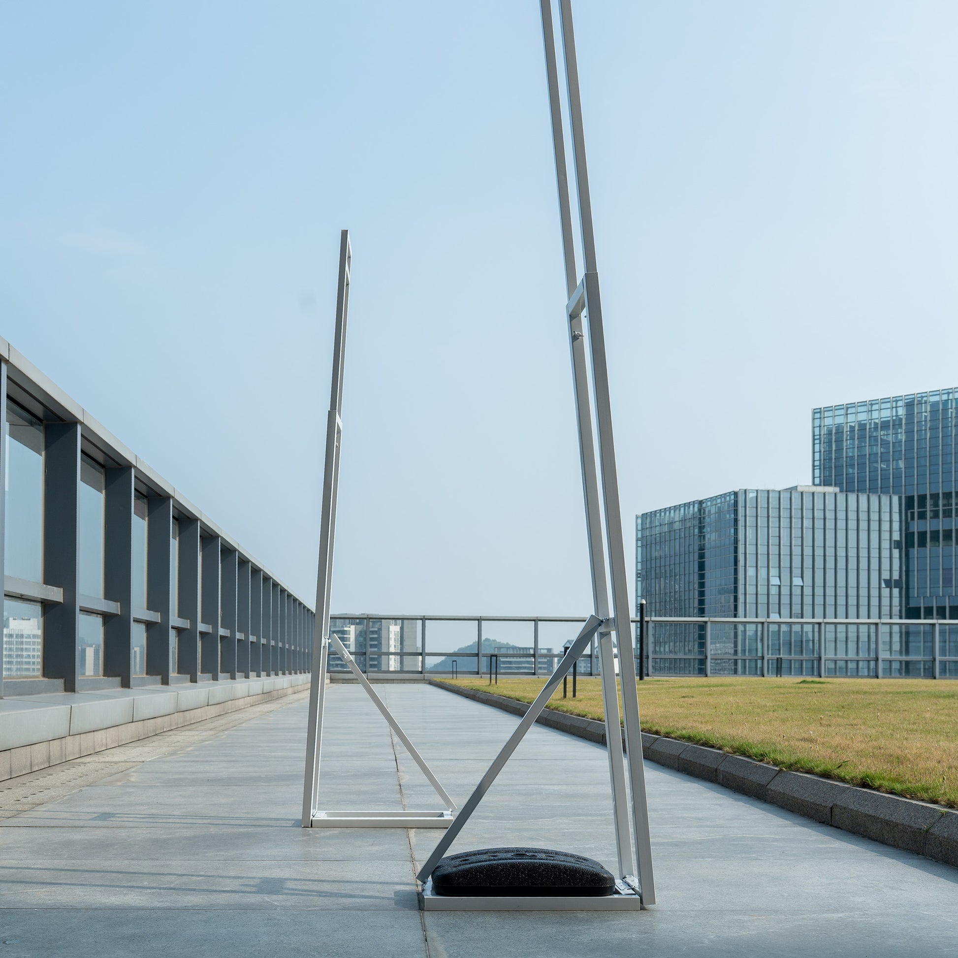 These backdrop stands are metallic structures positioned on an outdoor rooftop. They have a sleek, modern design with angled frames, giving them a contemporary aesthetic. The stands are equipped with black, weighted bases for stability, ensuring they can support floral or decorative backdrops for events. The urban cityscape in the background adds to the modern feel. 