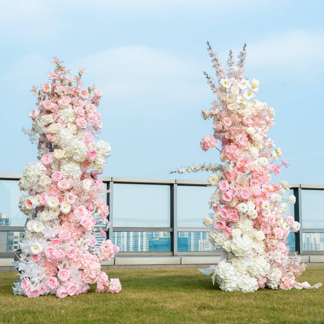 This backdrop features two tall floral arrangements placed outdoors on a rooftop with a cityscape in the background. The arrangements are composed of artificial flowers, including pink and white roses, white hydrangeas, and delicate pink blossoms that might be cherry - like flowers. The color palette is soft, with various shades of pink and white, creating a romantic and elegant style, perfect for wedding ceremonies or engagement photo shoots. 